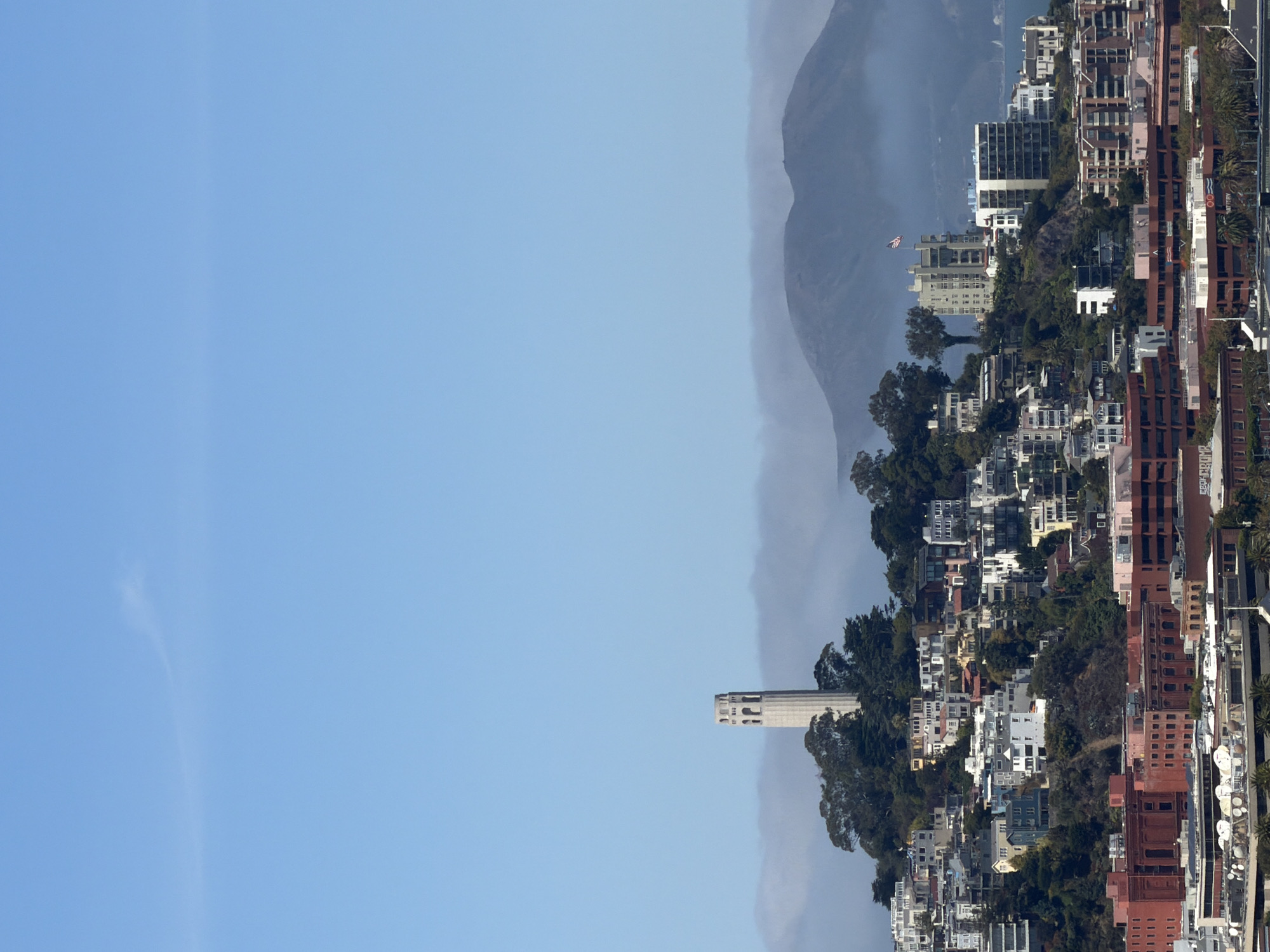 The image shows a view of San Francisco, California, United States of America, featuring a cityscape with a prominent tower. In the foreground, there are several multi-story buildings with red brick facades. Behind these, a densely populated hillside is covered with numerous houses and apartment buildings of various sizes and architectural styles, painted in a range of colors including white, blue, pink, and beige. Lush green trees are interspersed among the buildings, especially on the steeper slopes.
Dominating the mid-ground, on top of the highest point of the hill, stands the Coit Tower, a white cylindrical structure with a decorative open top. It is surrounded by trees and appears to be the focal point of the elevated section of the city. Further in the background, a range of mountains is visible, partially obscured by a layer of soft, white fog or clouds that stretches across the horizon. The sky above is a clear, pale blue with a faint, wispy cloud formation in the upper left quadrant. A U.S. flag flies from a building on the hillside to the right of Coit Tower. The overall scene suggests a bright, possibly cool day.