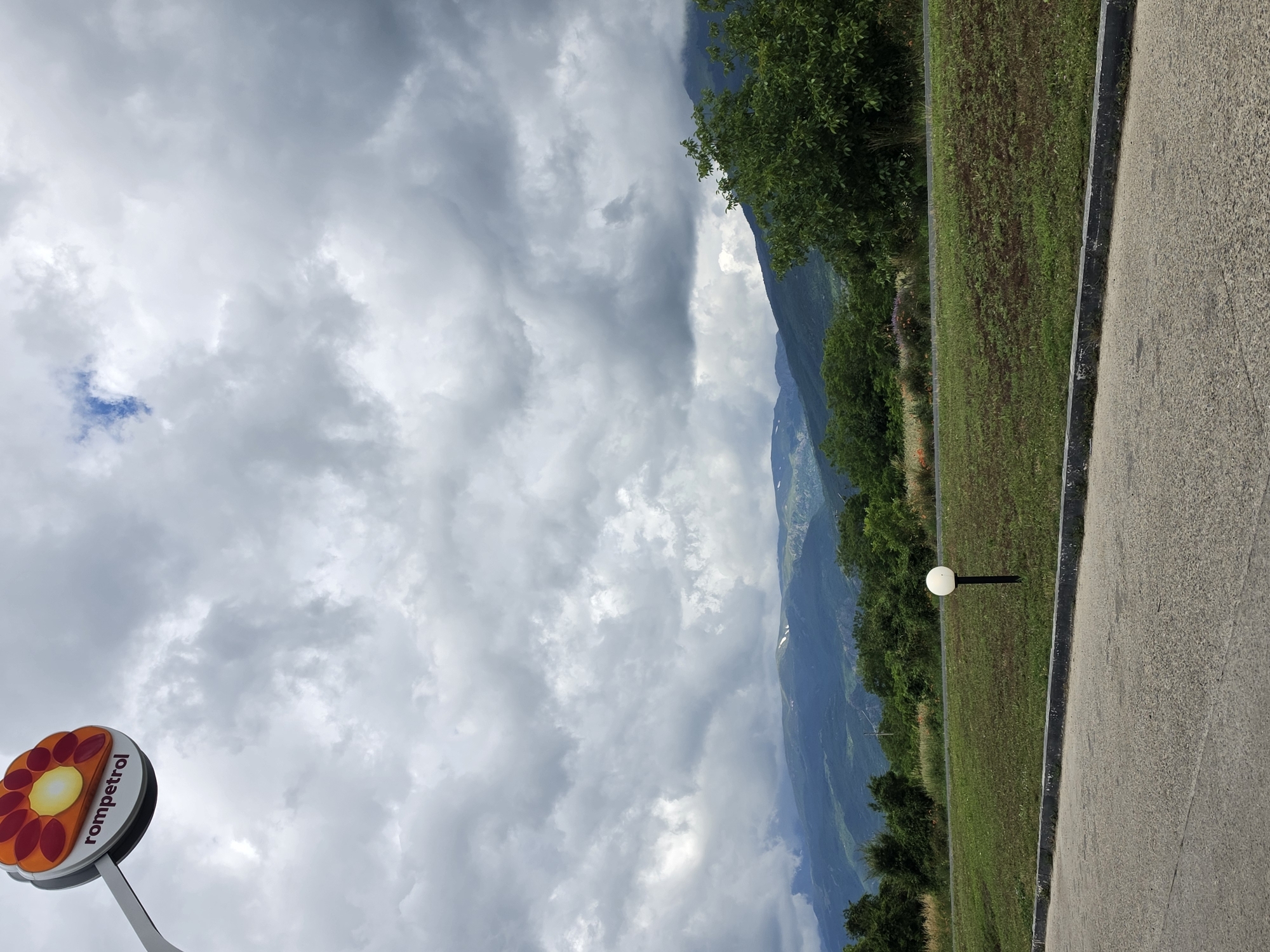 The image shows a cloudy sky over a mountainous landscape. In the foreground, there is a paved road and a grassy area with a single spherical lamp on a black pole. To the left, a sign with an orange and red flower design and the word "rompetrol" is visible. The background features lush green trees and rolling hills leading to more distant, darker mountains. The overall impression is a scenic view from a roadside location. The location is stated to be in the city of Tarnicheni, Bulgaria. There are no people or animals visible in the image, and no actions or interactions are taking place. The environment is natural, with a road and signage indicating a potentially commercial area like a gas station.