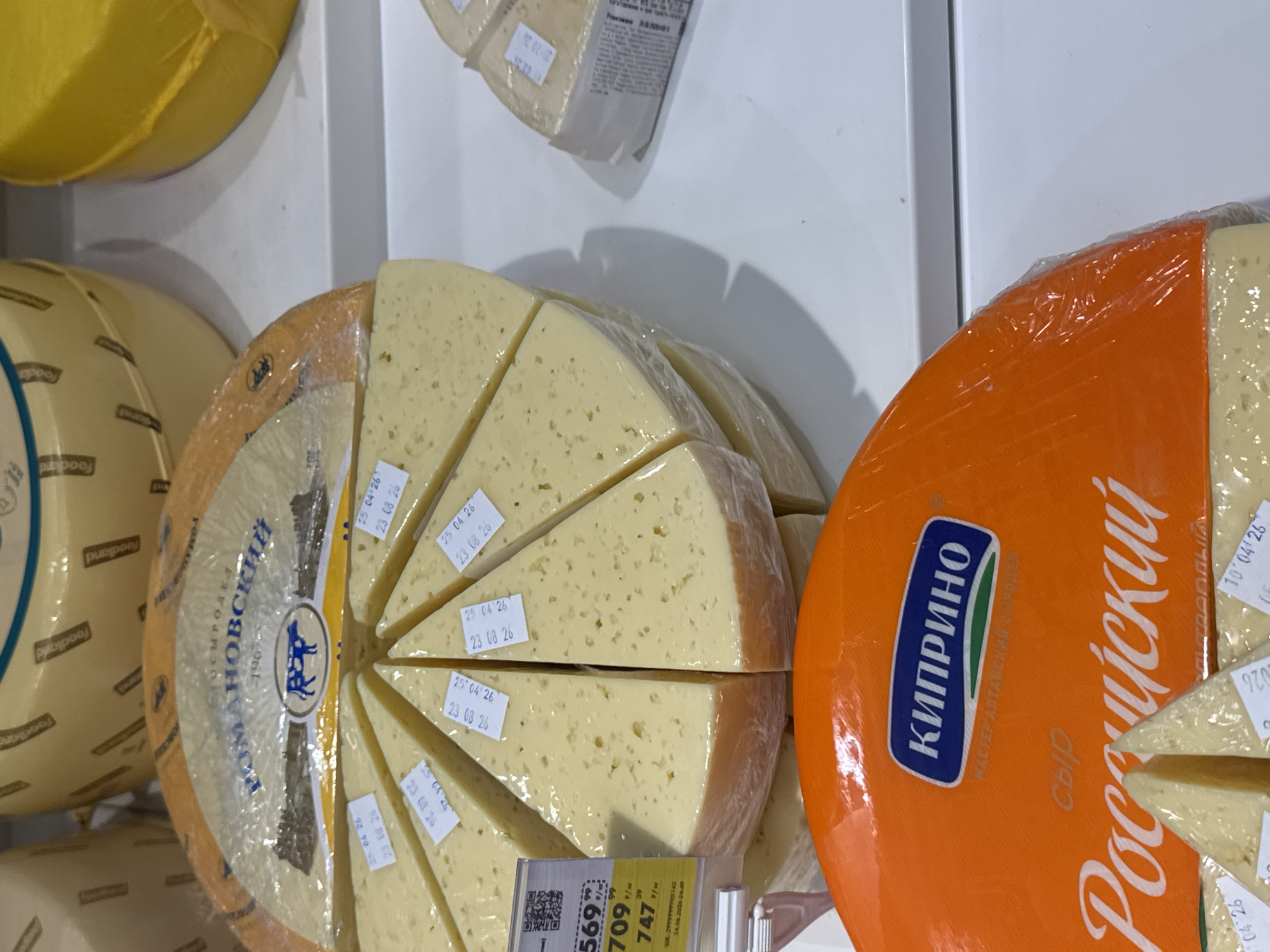 This media file shows a close-up, angled view of several wheels of cheese displayed in a retail setting, likely a grocery store or market. In the foreground, a large, bright orange wheel of cheese is prominent, with some slices cut from it revealing a pale yellow interior. White text in a stylized font is visible on the orange rind, including "КИПРИНО" and "Российский". To the left of the orange cheese wheel, a circular cheese, also sliced into wedges, is wrapped in plastic. This wheel has a label with Cyrillic text that appears to say "РОМАНОВСКИЙ" and features a blue graphic of a cow. Small white labels with dates, such as "25 04 26" and "23 08 26", are affixed to the cheese wedges. In the background, a portion of a large, yellow-wrapped cheese wheel is visible. The setting appears to be indoors with bright, even lighting. The focus is on the cheese products, with no people or specific time of day details evident. The overall impression is a display of dairy products for sale.