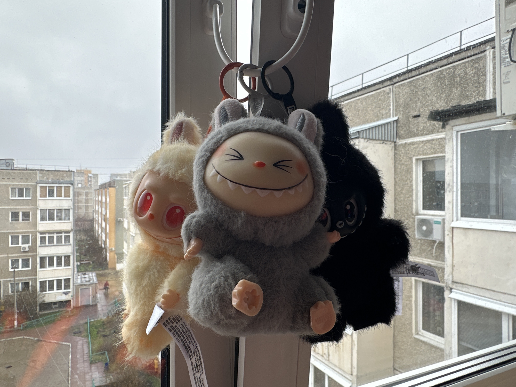 Three plush character keychains hang from white window handles, prominently positioned in the foreground of the image. From left to right, the keychains include a cream-colored creature with large red eyes, a central gray figure featuring a wide, toothy smile and squinted eyes, and a black fuzzy creature with dark eyes, partially obscured. All three appear to be stylized monster or creature designs.
Beyond the window, a gray and overcast sky blankets the scene, with faint specks in the air suggesting light precipitation like snow or sleet. The outdoor environment depicts a residential area in an urban or suburban setting, likely Chaikovsky, Russia. Multiple multi-story apartment buildings, characteristic of post-Soviet architecture with their concrete facades and repeating window patterns, are visible. A courtyard with bare trees, paved pathways, and some patches of grass lies between the buildings. The overall impression is that of a cold, dreary daytime scene.
Tags hanging from the keychains display visible text. OCR identifies phrases such as "THE MONSTERS" in English, along with Chinese characters, a phone number (400-023-2233), and address information, likely detailing the manufacturer or brand. The bright, smiling face of the central gray plush provides a cheerful contrast to the muted outdoor weather.