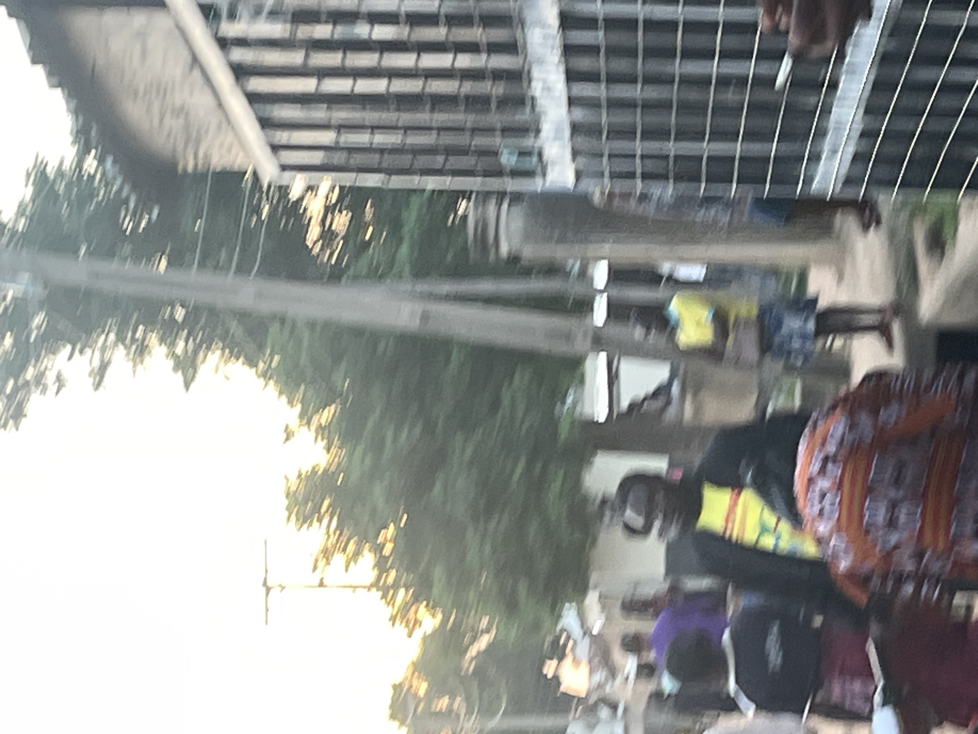 The image depicts a street scene in Jalingo, Nigeria, characterized by a bright, overcast sky and visible power lines and poles. In the foreground, several individuals are present. A man wearing a yellow t-shirt with horizontal red and yellow stripes and a dark jacket is prominently featured. To his left, a person in a black shirt and red checkered skirt is partially visible. Behind them, other figures, including one in a purple shirt, are present, though their features are indistinct due to motion blur.
To the right of the scene, a dark metal gate with a grid pattern is visible. Beyond the gate, a stone building with an arched structure is partially seen. In the background, lush green trees and other structures line the street. The overall impression is of a busy outdoor gathering or thoroughfare.