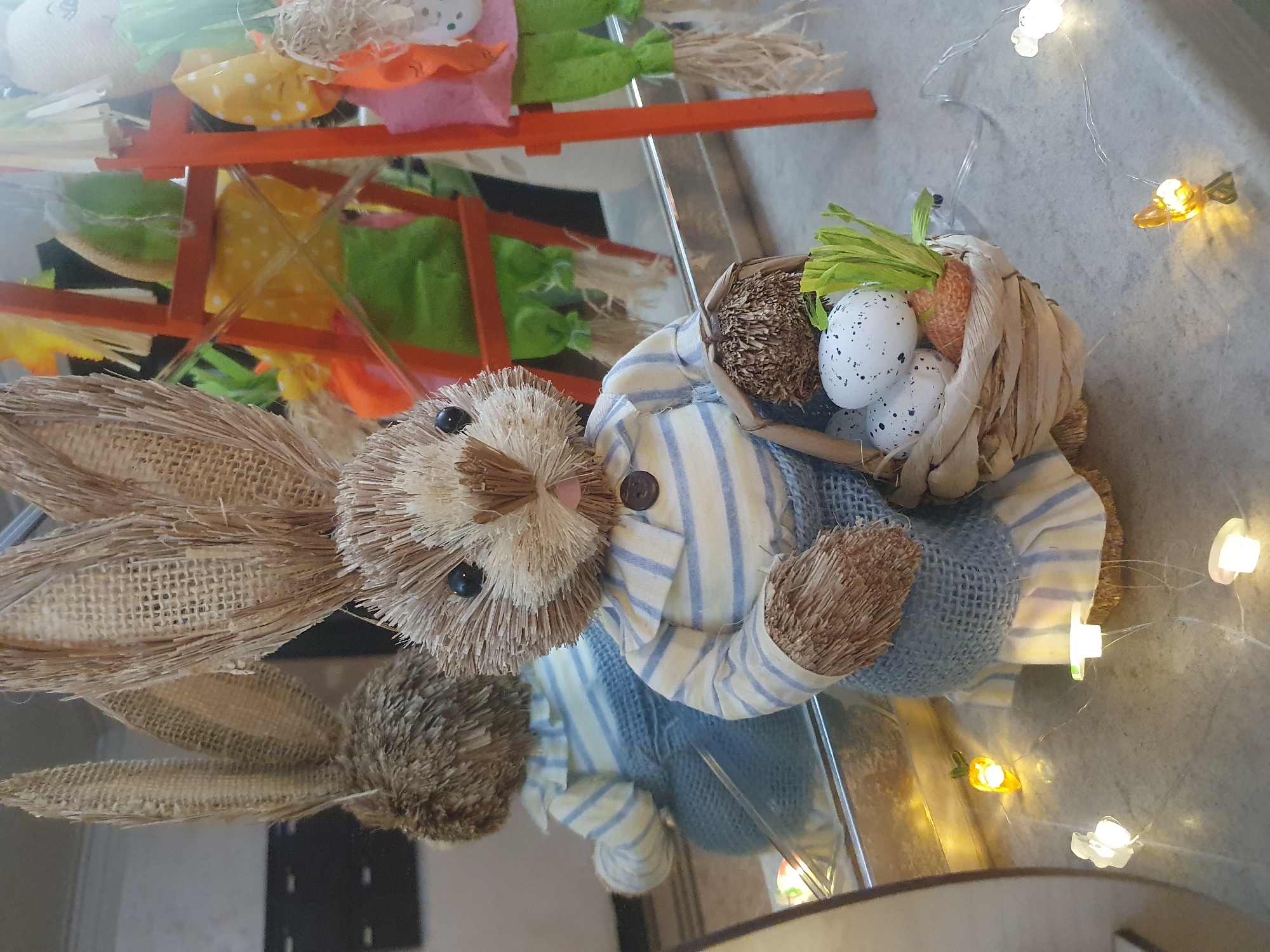 The image shows a straw-like rabbit figurine wearing a blue-striped shirt and overalls. It is sitting next to a woven basket containing straw, speckled eggs, and a toy carrot with green leaves. A miniature wooden stand with colorful decorations is situated behind the rabbit. The scene is illuminated by string lights featuring flower and carrot-shaped bulbs. The reflection of the rabbit figurine is visible on a glossy surface. The location, Plovdiv, Bulgaria, cannot be determined from the image alone.