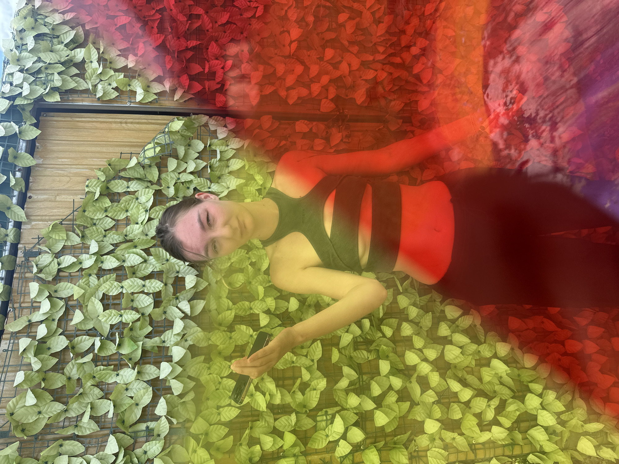 A young female subject stands centrally in a full-length portrait against a dual-toned artificial foliage background in Nghia Lo Ward, Vietnam. She has light skin, dark hair pulled back, and maintains direct eye contact. Dressed in a dark halter-neck crop top with several horizontal midriff cutouts and dark, form-fitting pants, she holds a black smartphone with a gold/bronze frame horizontally in her open right hand. The background features light green artificial leaves on a grid to the left and red artificial leaves on a grid to the right, with a light brown wooden panel visible between them centrally in the upper frame. The entire scene is heavily overlaid with a color gradient, presenting a strong yellow hue on the left transitioning through orange and red to deeper red and purple tones on the right.