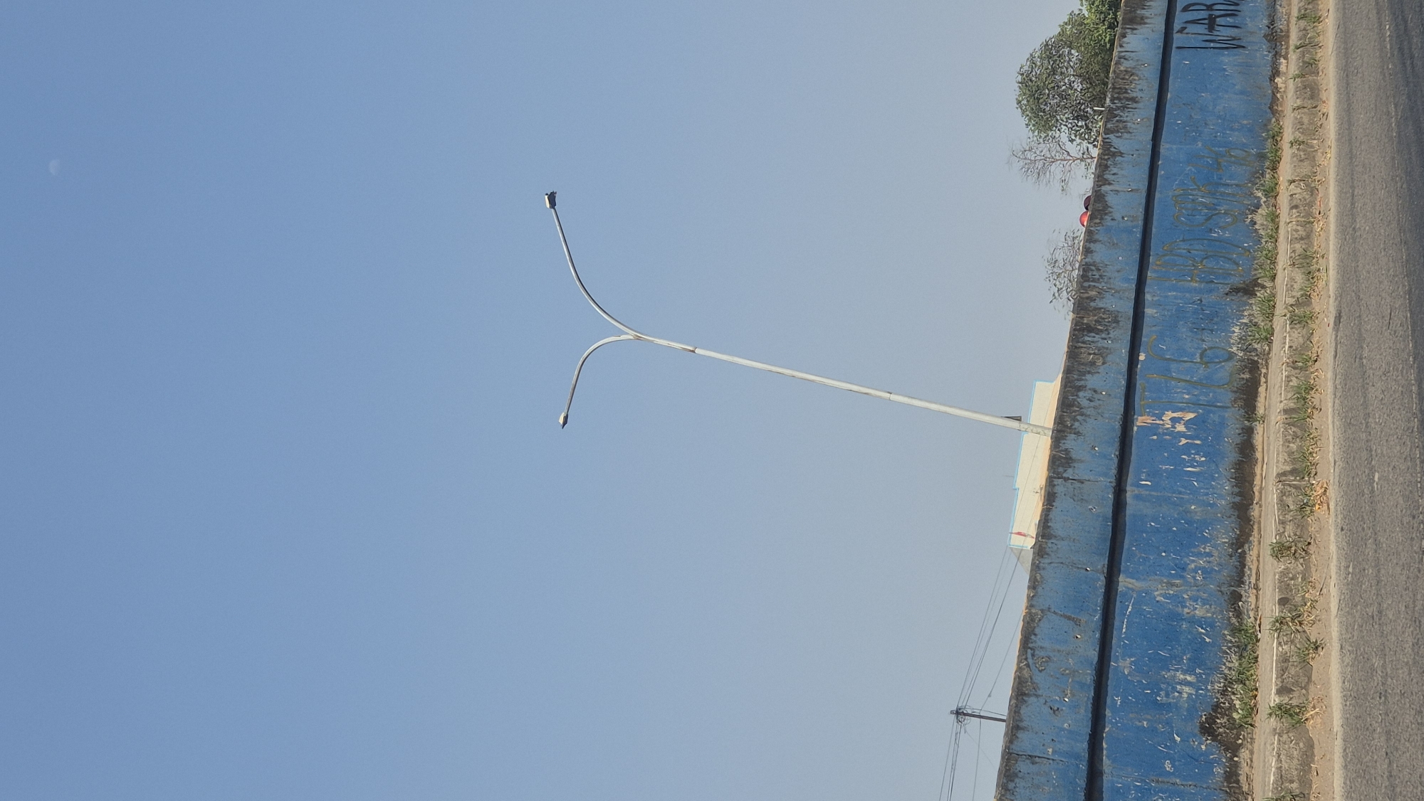 A street lamp with two arms extends into a clear blue sky, positioned above a blue concrete barrier in Karawang, Indonesia. The barrier, showing signs of age and graffiti, runs horizontally across the mid-ground. Small patches of green vegetation grow along the base of the barrier and on the sidewalk. The sky is bright and cloudless, with a faint, small moon visible in the upper right corner. The scene appears to be an outdoor urban environment, possibly near a road given the visible pavement in the foreground.