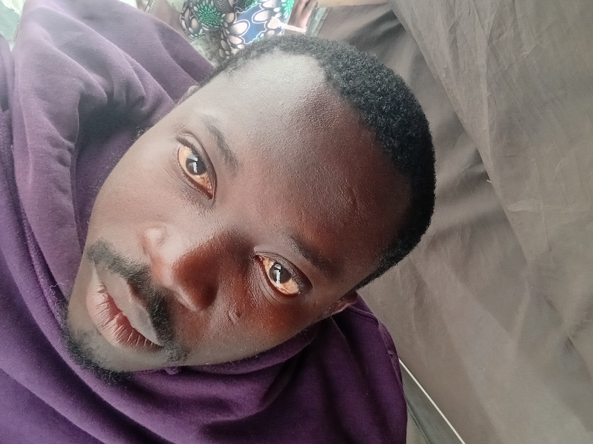 A close-up, high-angle, selfie-style photograph shows a dark-skinned Black man from the chest up, wearing a purple hooded sweatshirt that is pulled up around his neck and chin. The man has short, dark, curly hair, a mustache, and a goatee. His eyes are open, and his irises appear yellowish. The background is a neutral, draped fabric, possibly a tent or tarp, in a light gray color. There is a hint of a colorful patterned fabric on the left side of the image, partially obscured by the man's sweatshirt. The lighting is diffused, suggesting an indoor or overcast outdoor setting. The image is composed to focus on the man's face, with the top of his head and forehead prominently featured.