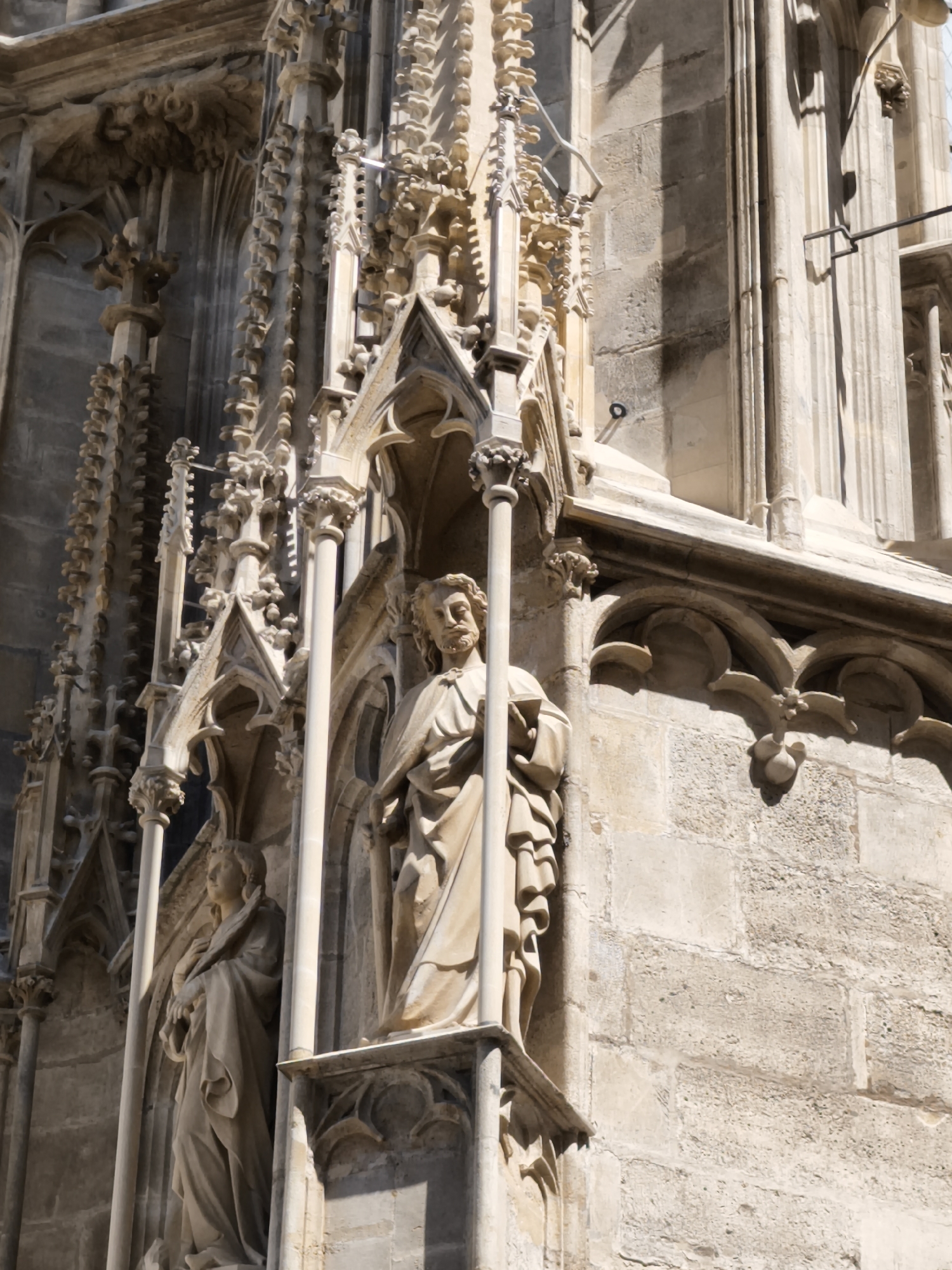 This is a detailed, close-up shot of the exterior of St. Stephen's Cathedral (Stephansdom) in Vienna, Austria. The photograph focuses on the intricate Gothic architecture of the cathedral's facade, specifically highlighting two stone statues positioned within alcoves. The statue on the right depicts a male figure with a beard, wearing a draped robe and holding an object in his hands, possibly a book. The statue on the left is of a female figure, also in a draped robe, with her hands clasped. Both statues are weathered and show signs of age. The surrounding stonework is highly ornate, featuring pointed arches, carved decorative elements, and vertical finials. Sunlight casts strong shadows on the textured stone, emphasizing the depth and complexity of the architectural details. The overall scene conveys a sense of historical grandeur and artistic craftsmanship.