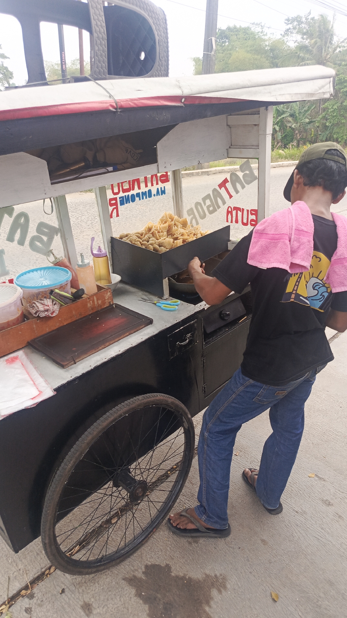 A person wearing a dark t-shirt, blue jeans, and flip-flops stands beside a food cart in Parung Panjang, Indonesia. The person has a pink towel draped around their neck and is reaching towards a container of fried food on the cart. The food cart is black and has a large spoked wheel on its left side. There are various containers and bottles on the cart's surface, including a blue-lidded container and two squeeze bottles. A sign with text, partially obscured and reversed, is visible on the cart. The background shows a road and vegetation, suggesting an outdoor setting.