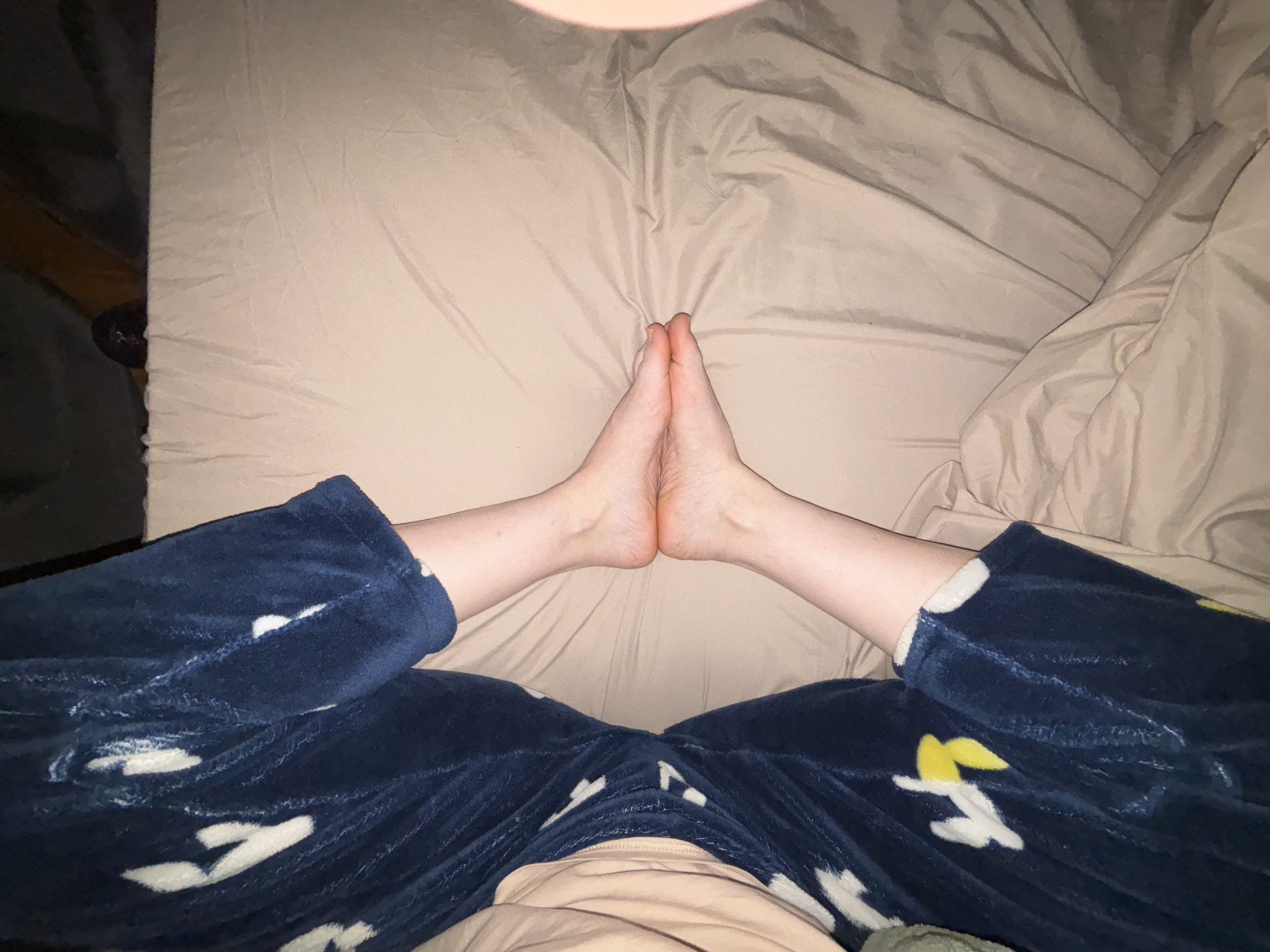 A person lies on a bed covered with a tan-colored sheet and pillow. The person is wearing blue pajama pants with a white pattern. The person's bare feet are touching each other at the center of the bed. The location and city are unknown.