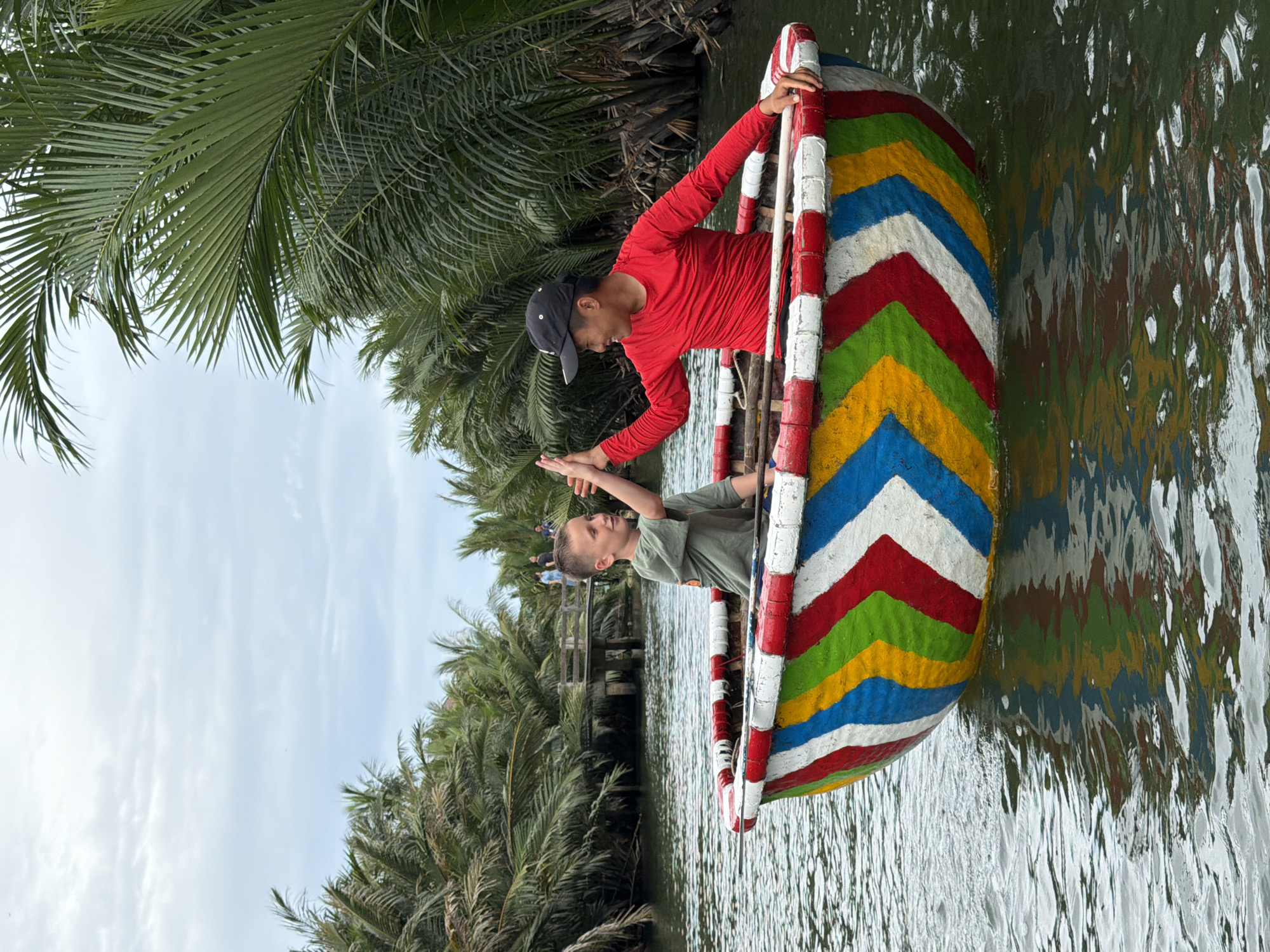 Two individuals, a young boy and a man, are in a brightly painted round boat, often called a basket boat, on a calm river. The boat is decorated with a chevron pattern of red, white, blue, yellow, and green. The boy, wearing a green t-shirt, is on the left, and the man, in a red long-sleeved shirt and a black baseball cap, is on the right. They are both smiling and giving each other a high-five. The man is holding an oar. The boat is surrounded by lush green palm trees and vegetation, characteristic of the coconut palm groves found in the Hoi An area of Vietnam, specifically in the Cam Thanh coconut village. A wooden bridge and a few other people are visible in the background across the river. The water reflects the colorful boat and the surrounding greenery.