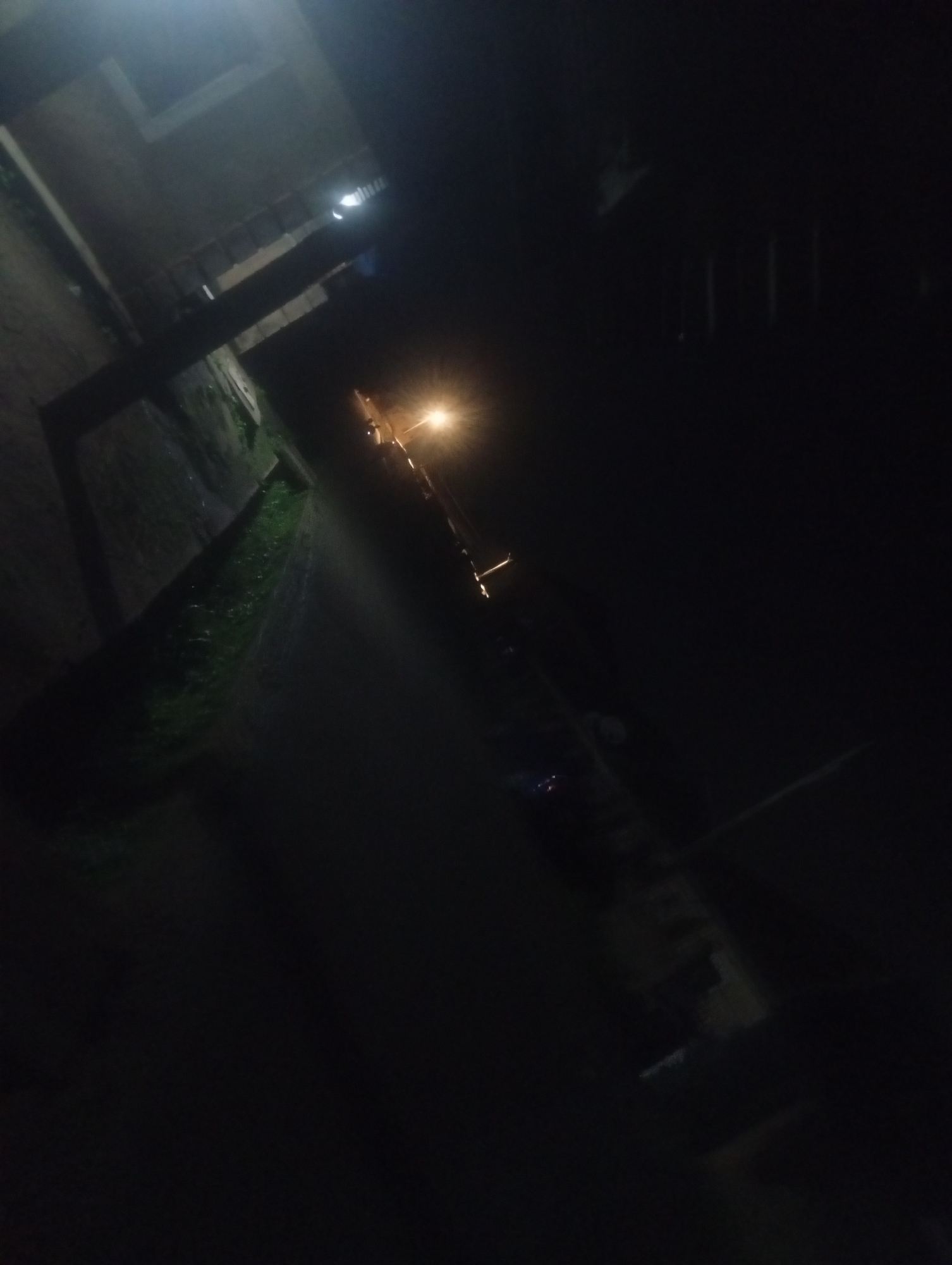 The image captures a dimly lit street scene at night in Bukuru, Nigeria. A street lamp illuminates a portion of a narrow, paved road. To the left of the road, a concrete wall runs parallel, with a strip of overgrown green grass or weeds growing along its base. Beyond the wall, structures are vaguely visible in the darkness, with hints of windows or openings. The scene appears deserted, with no people or vehicles present. The overall impression is one of a quiet, somewhat neglected urban area after dark, reliant on the single street light for visibility.