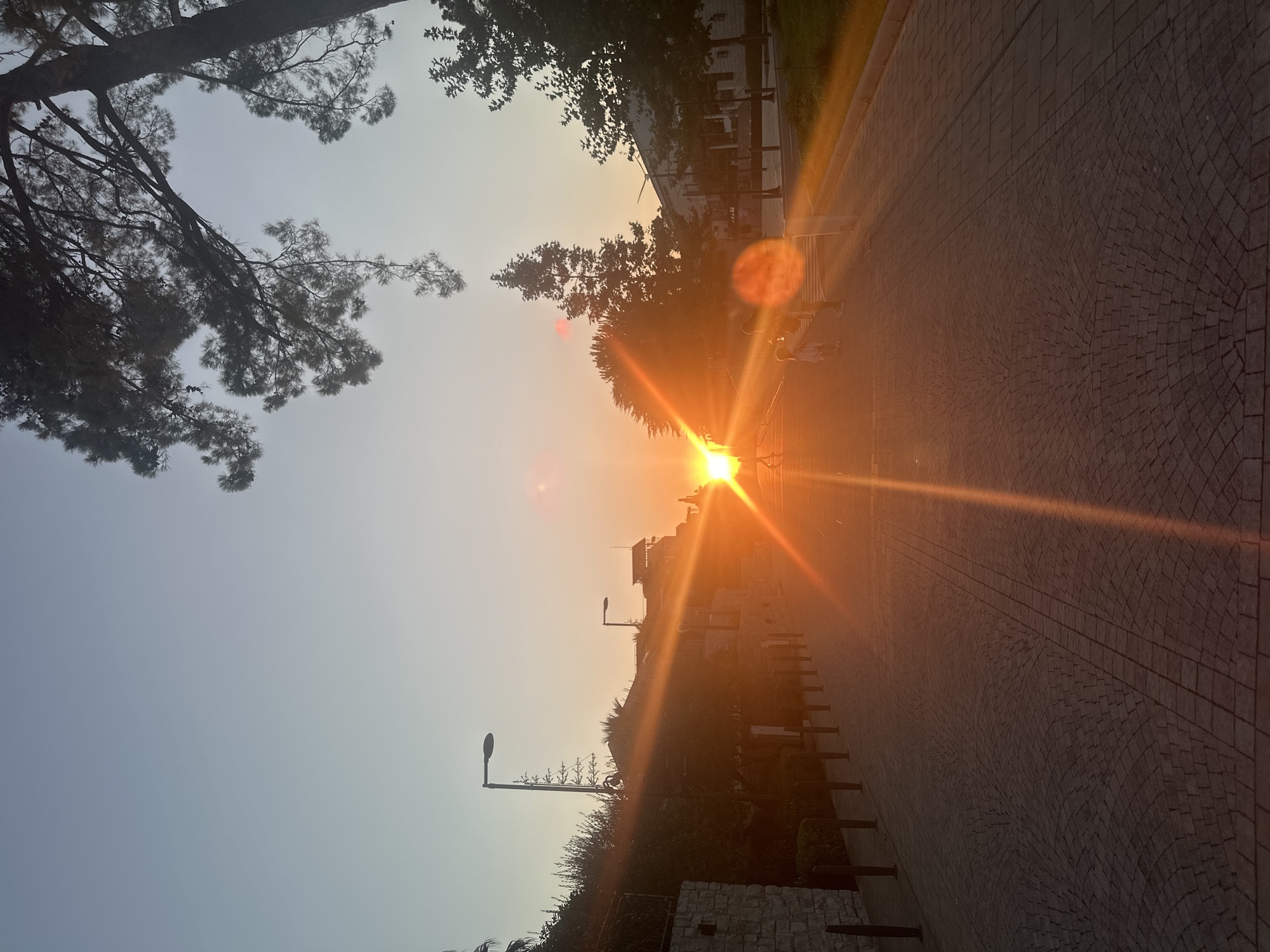 The image depicts a sunset over a paved pathway in Kumbor, Montenegro. The sun is low on the horizon, casting a strong orange glow and lens flares across the scene. The pathway is made of dark cobblestones arranged in a fan pattern. On the left side of the pathway, there is a stone wall and vegetation. On the right, there are trees and modern buildings, some of which appear to be residential or commercial. Two lampposts are visible on the left side of the pathway, one closer and one further away. In the middle ground, a couple of individuals, one adult and one child, are walking on the pathway. The sky transitions from a pale blue near the top to a warm orange and yellow near the horizon. Pine trees are visible in the upper right corner, with their branches silhouetted against the sky. The overall atmosphere is serene, with the setting sun illuminating the scene.
