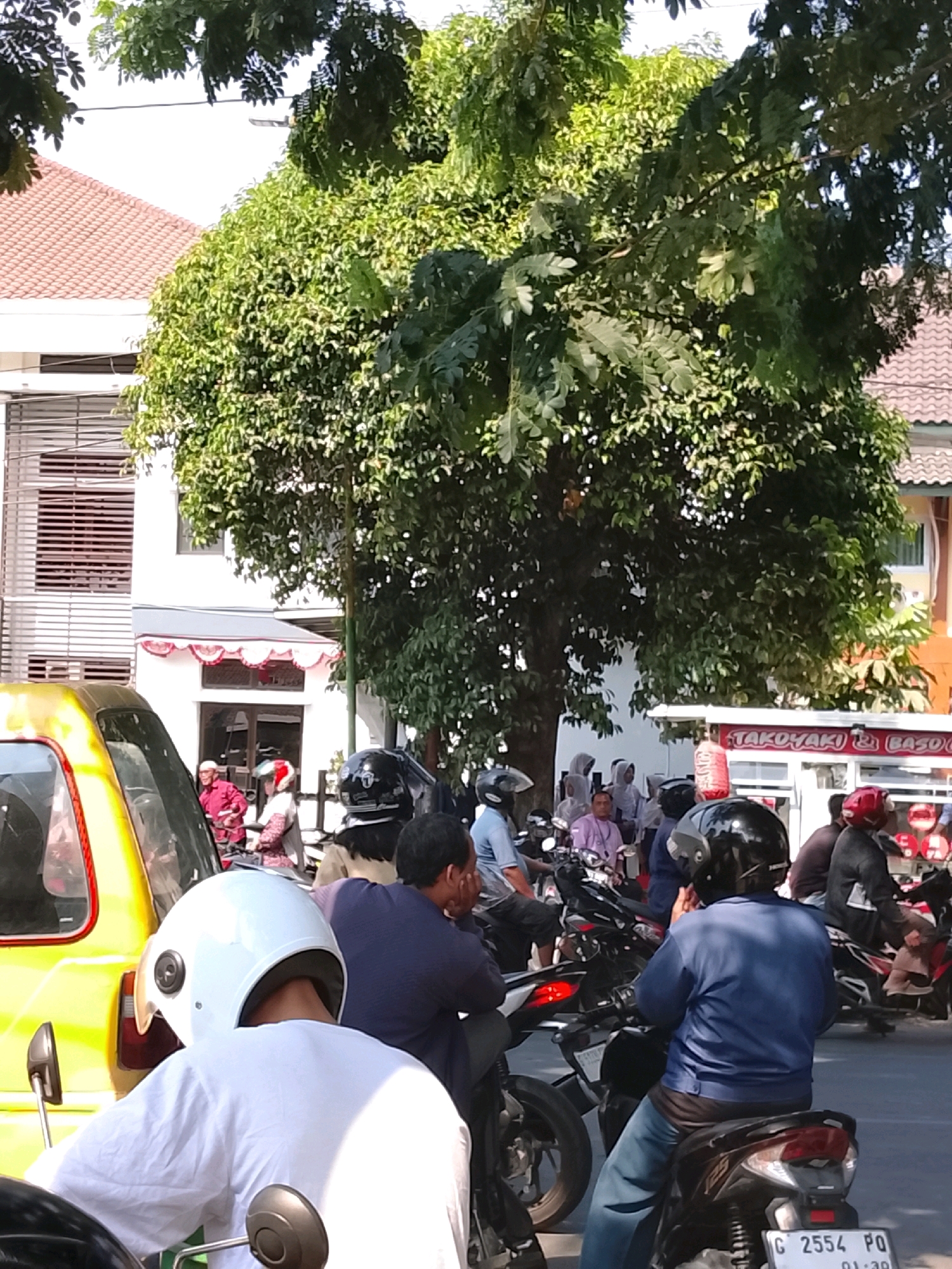 The image captures a street scene in Slawi, Indonesia, likely a commercial area with a building featuring a red-tiled roof and a white facade with horizontal blinds. A large, leafy green tree dominates the upper half of the frame. In the foreground and middle ground, a yellow vehicle, possibly a van or car, is visible on the left. The primary focus is on a group of people on motorcycles. Several individuals are wearing helmets, including one in the foreground wearing a white helmet and a white shirt, and another in the middle ground with a black helmet and a blue shirt. Other motorcyclists are also present, some with helmets. In the background, a food stall with the text "TAKOKYAKI & BASO" is partially visible. The overall impression is of a busy street with people engaged in everyday activities, likely during the daytime given the bright lighting.