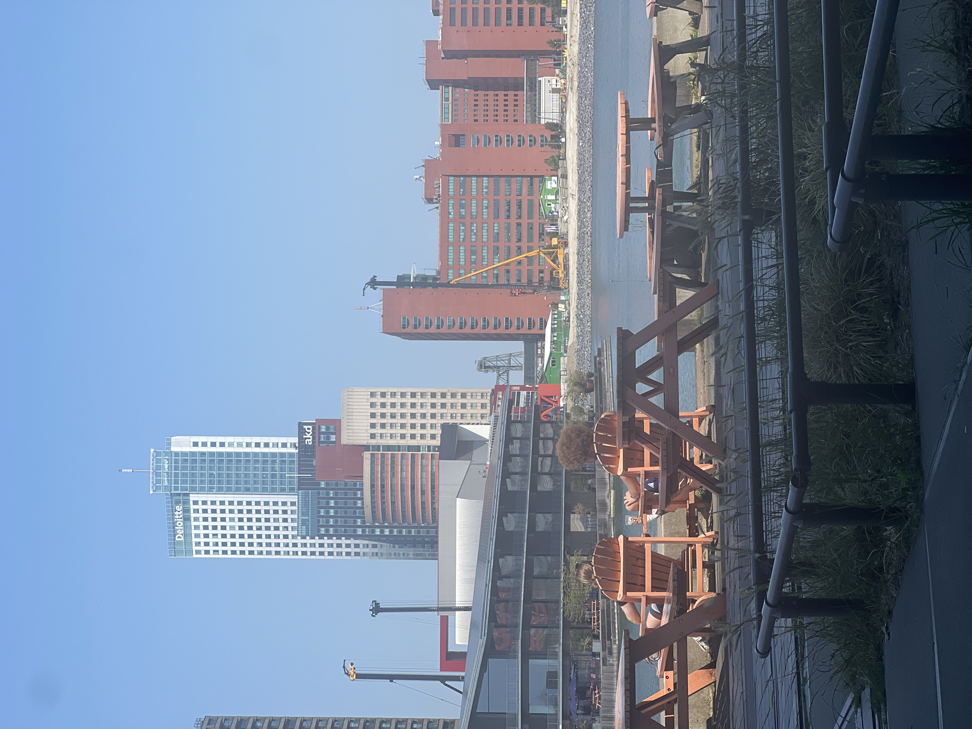 This is a vertically oriented photograph taken outdoors, likely on a sunny day given the clear blue sky and strong shadows. The scene is a waterfront area in Rotterdam, Netherlands, featuring a cityscape dominated by modern architecture.
In the foreground, there are several wooden Adirondack chairs and picnic tables set up along a paved walkway, with some grass and shrubbery bordering the area. Two individuals are partially visible sitting in the Adirondack chairs, with one person's legs and feet clearly visible, wearing shorts and sneakers. The presence of these seating areas suggests a public space or an outdoor dining/lounge area.
Behind the seating, a body of water is visible, and beyond that, a dense collection of buildings forms the Rotterdam skyline. These buildings are varied in design, with some being tall, modern skyscrapers and others appearing to be older, red-brick structures. Notable buildings include a prominent white skyscraper with the "Deloitte" logo and another building with the "akd" logo. Cranes are also visible among the buildings, indicating ongoing construction or development in the area.
The overall impression is one of a vibrant urban environment situated along the water, with public amenities for relaxation and enjoyment of the city views. The clear sky suggests it's daytime, likely late morning or afternoon.