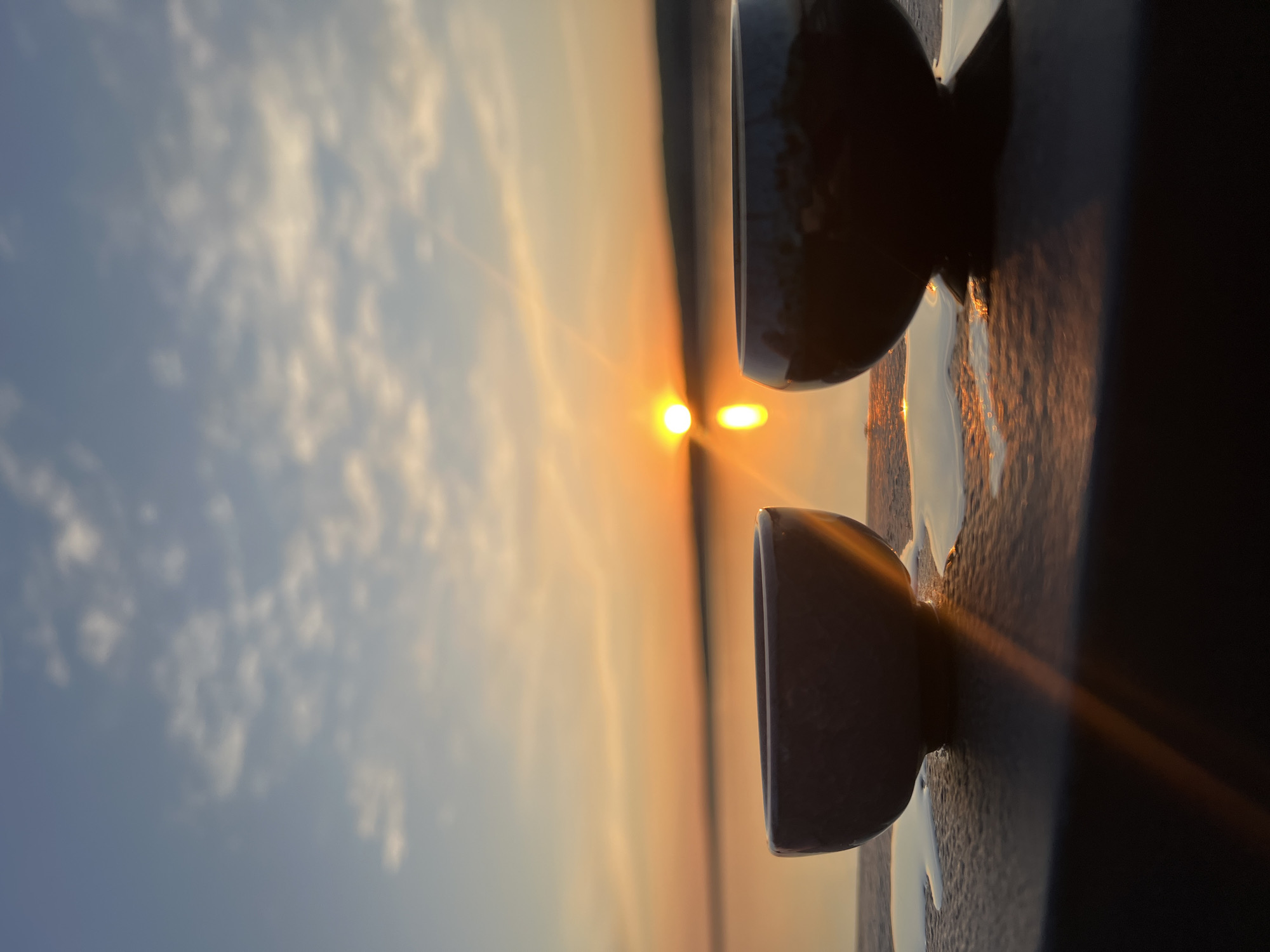 The image shows a horizontal view of a sunset with two small bowls placed on a dark surface, likely a ledge or wall. The sky is a gradient of blues and oranges, with the sun shining brightly on the horizon. There are some faint clouds in the sky. The surface on which the bowls sit is textured and dark, reflecting some of the light from the sunset. The bowls are a light grey/beige color and are placed on either side of the horizon. Based on the location request of the city of Chaikovsky, Russia, this photo was likely taken in that location.