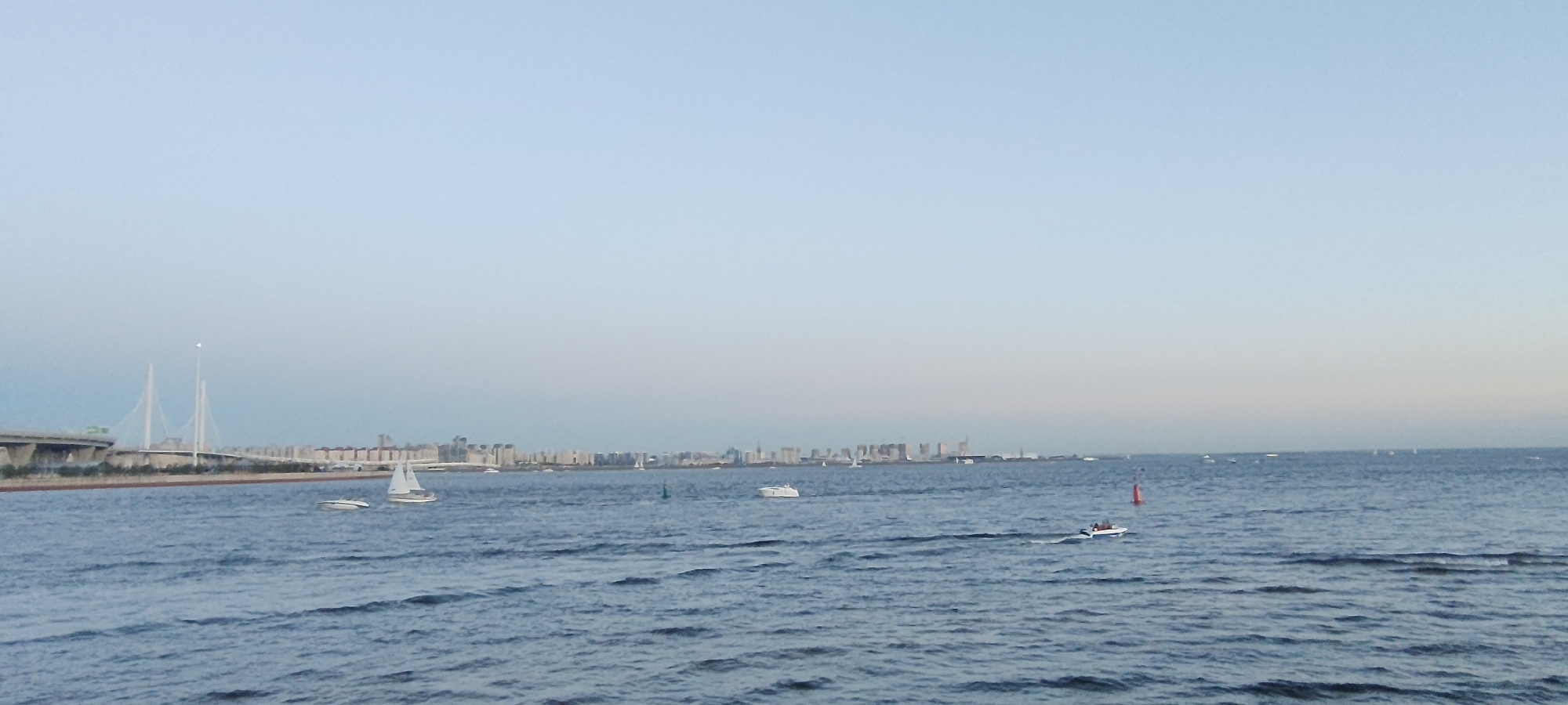 This is a horizontal, eye-level shot of a body of water in Saint Petersburg, Russia, with the city skyline in the background. The sky is a pale blue with a gradient of lighter blue towards the horizon, indicating either dawn or dusk.
In the foreground, the water is a deep blue with visible ripples and small waves. Several boats are scattered across the water. On the left side, a white sailboat with two sails is visible. To its right, a white motorboat is moving. Further to the right, another white sailboat is sailing. In the mid-ground, a red buoy is visible. Towards the right, a small white motorboat with people on board is moving, leaving a wake.
In the background, the Saint Petersburg skyline stretches horizontally across the image. The most prominent structures are the twin towers of the cable-stayed bridge, also known as the Western High-Speed Diameter, on the left. Further to the right, a long line of buildings forms the city's silhouette, with some taller structures interspersed. The overall impression is one of a calm, expansive scene with the city as a backdrop.