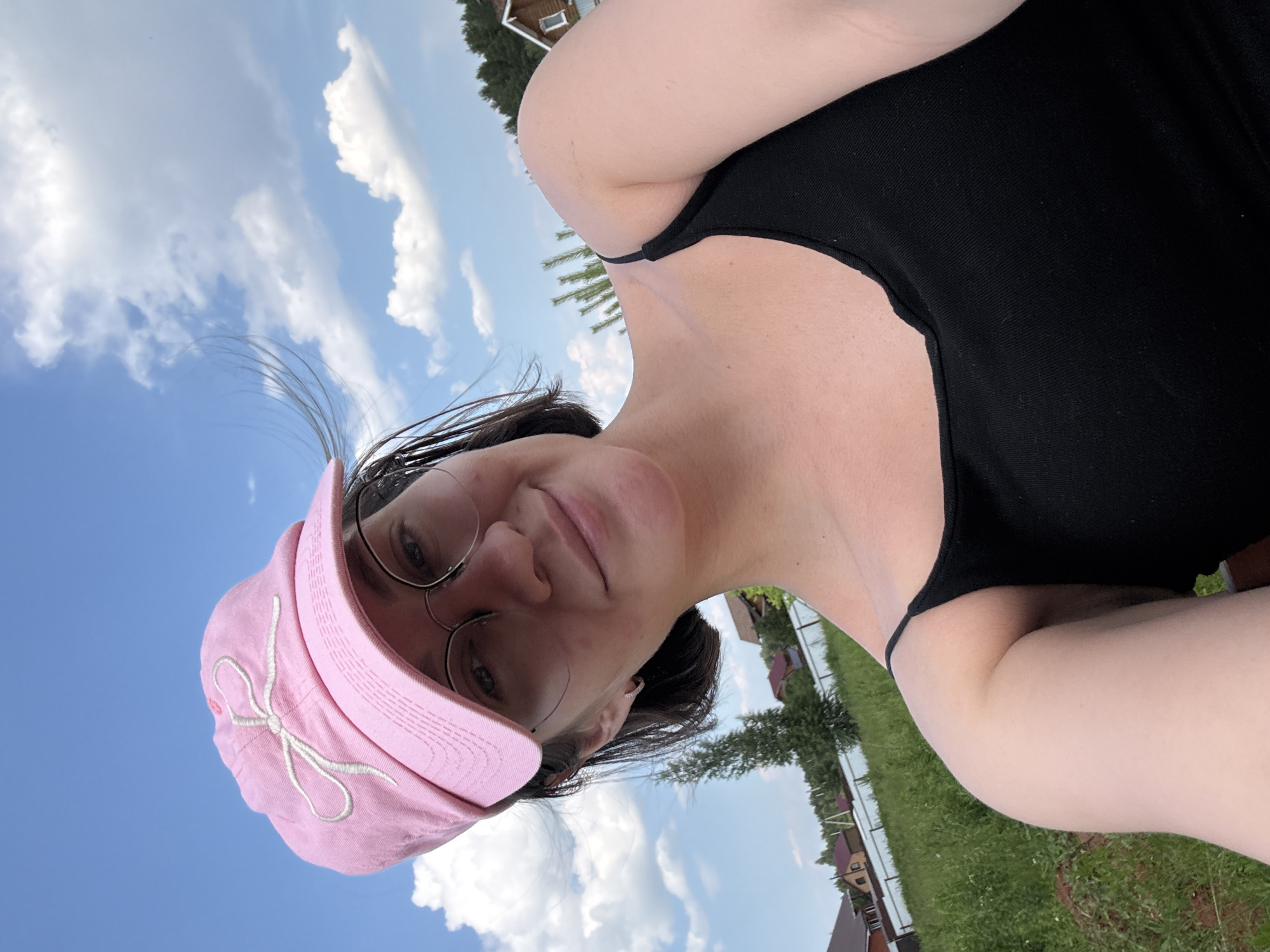 A young woman with short, dark hair is taking a selfie outdoors on a sunny day. She is wearing a pink baseball cap with a white embroidered bow and round-rimmed glasses. Her expression is a slight smile. She is wearing a black tank top. The background shows a blue sky with white clouds, green grass, a white fence, and several houses with red roofs, suggesting a suburban or rural setting. Trees and foliage are also visible. The image is likely taken in the city of Dokshitsy, Belarus, not Doksha, Russia.