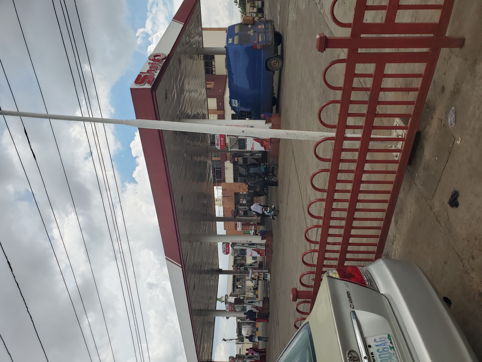 The image depicts a busy gas station in Jos, Nigeria, under a partly cloudy sky. The scene is filled with various vehicles, including a blue van parked under the canopy, several motorcycles, and cars, one of which, a silver Toyota Camry, is partially visible in the foreground. People are seen interacting with the vehicles and pumps, suggesting an active refueling or service operation.
The gas station canopy is prominently featured, with the "Shell" logo visible on some of the pumps and signage. The red metal barrier in the foreground adds a distinct visual element to the scene. The overall atmosphere appears to be one of everyday commerce and activity. The presence of power lines overhead suggests an urban or semi-urban environment. The license plate of the Toyota Camry shows "306GAG" and "ABUJA". The weather appears to be overcast with patches of blue sky, indicating it could be late morning or afternoon.