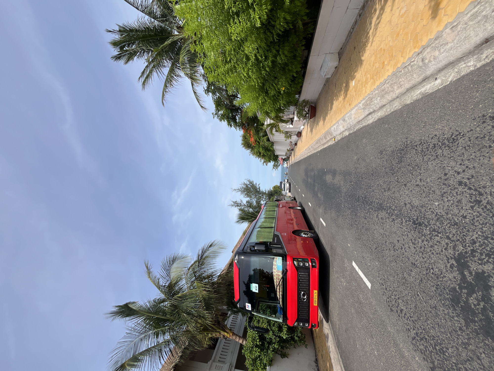 A red tour bus is parked on the side of a paved road in Mũi Né, Vietnam. The bus is angled towards the camera, with its front facing left. The road is asphalt with white lane markings. To the left of the road, there is a white building with palm trees and lush green bushes. To the right of the road, there is a yellow tiled sidewalk with a low white wall and more green foliage. In the distance, other vehicles and people can be seen. The sky is blue with scattered clouds, and the weather appears to be sunny.
