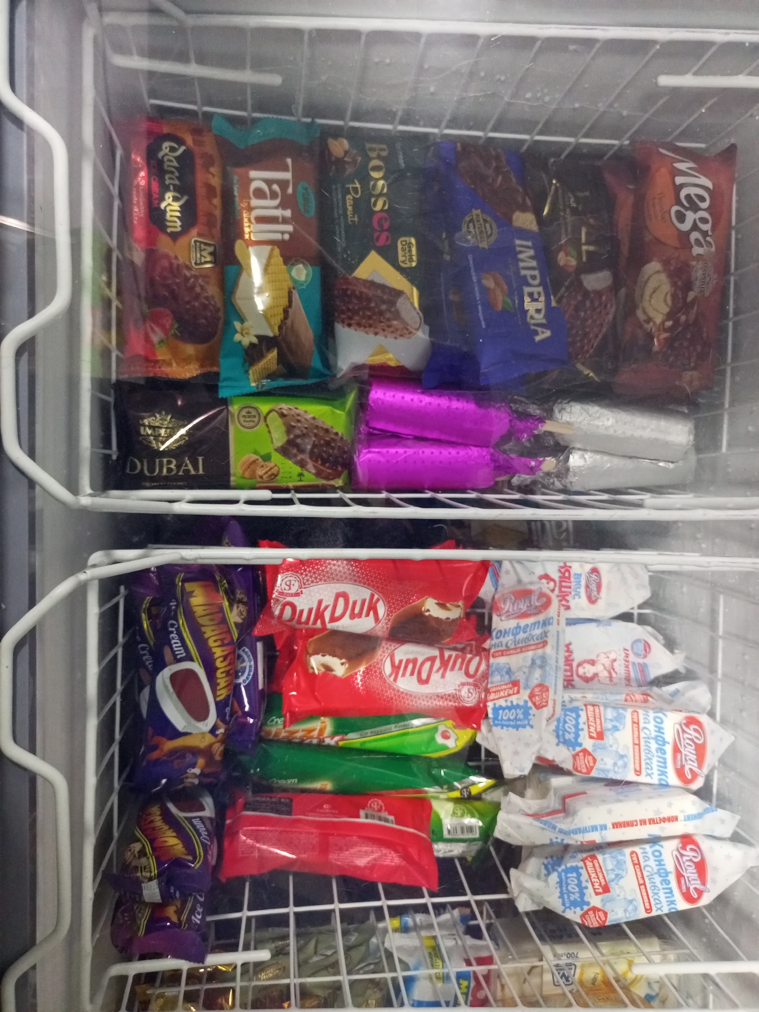 This is a medium shot, eye-level, indoor photograph of a freezer display case stocked with various ice cream products. The freezer is divided into two shelves. The top shelf contains an assortment of individually wrapped ice cream bars and cones. Brands visible include "Mega," "Bosses," "Tatti," "Qara-Qum," "Imperia," and "Dubai." Some wrappers are colorful and feature images of ice cream.
The bottom shelf also contains a variety of ice cream products. Prominently displayed are red and green wrappers, some with the "DukDuk" brand. There are also white wrappers with blue text, some appearing to be ice cream bars or popsicles, with the word "Royal" visible. Two bright pink wrappers are also present on the top shelf, each containing what appears to be a popsicle.
The overall environment suggests a retail setting, likely a small shop or convenience store. The freezer shelves are made of white metal wire. The ice cream packaging is diverse in design and color, indicating a range of flavors and brands. The lighting is artificial and appears to be coming from above. The location is identified as Kitab, Uzbekistan. There are no people or animals visible in the image.