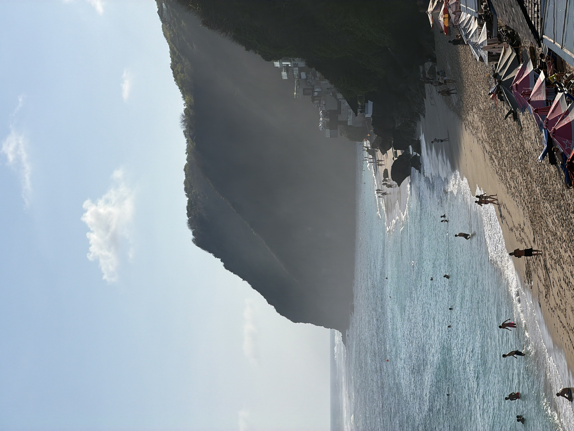 This image depicts a sun-drenched beach scene in Kuta Selatan, Indonesia, likely near a cliffside resort or development. The ocean, with its gentle waves, occupies the left side of the frame, transitioning to a sandy beach on the right. Numerous individuals are scattered across the scene. Several are wading in the shallow water, while others are walking along the shoreline or standing on the sand. Beach umbrellas and lounge chairs are visible on the sand, suggesting a recreational area. In the background, a dramatic cliff face covered in vegetation rises, with structures that appear to be part of a resort or dwellings built into the hillside. The sky is a clear blue with a few wispy clouds, and the overall atmosphere is one of a tranquil beach day.