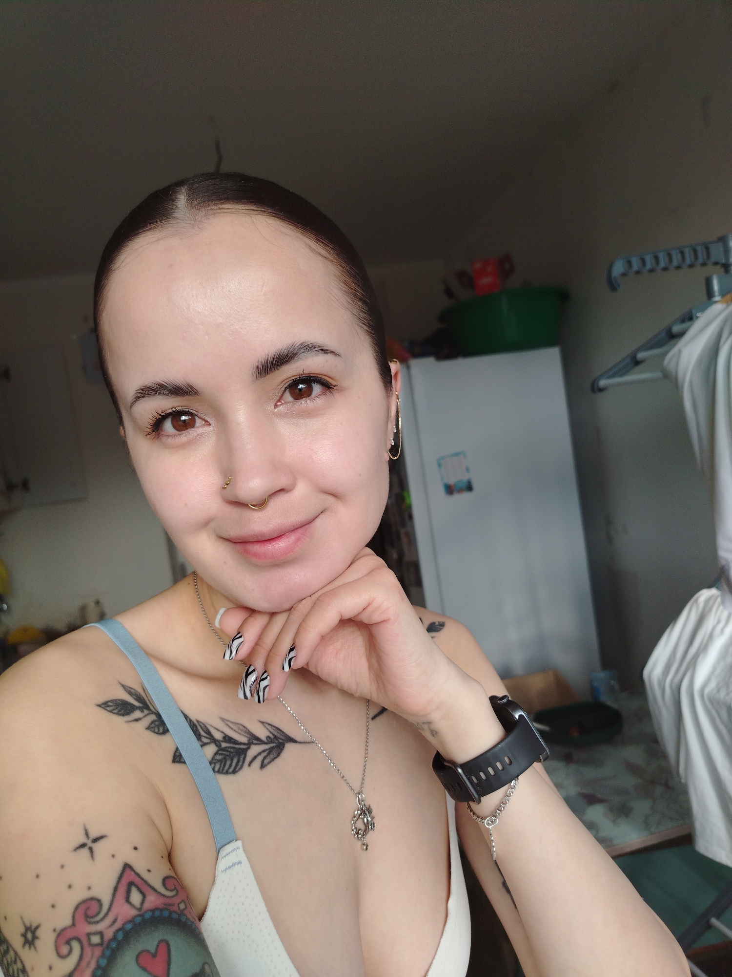 A young woman is featured in a close-up selfie, smiling gently with her right hand resting under her chin. She has dark hair pulled back, revealing her forehead, and well-defined eyebrows framing her warm brown eyes. A gold hoop nose piercing adorns her nose, and a delicate gold chain earring hangs from her left ear. Her nails are painted with a distinctive black and white zebra print design.
She is wearing a light-colored bra with a pale blue strap visible. Her skin is adorned with several tattoos: a leafy branch design on her left shoulder/chest, and a larger, colorful tattoo featuring geometric shapes, stars, and a heart on her left upper arm. A black digital smartwatch and a silver chain bracelet are visible on her right wrist, and she wears a silver chain necklace with a pendant.
The indoor setting appears to be a casual home environment, possibly a kitchen or utility room, illuminated by natural daylight from the left. In the background, a white refrigerator is visible on the right, topped with a bright green plastic basin. Part of a clothes drying rack with white laundry is also discernible. The woman's expression is calm and friendly. No text is visible in the image. The scene suggests a relaxed moment in a domestic setting, consistent with a home in Noviy, Russia.