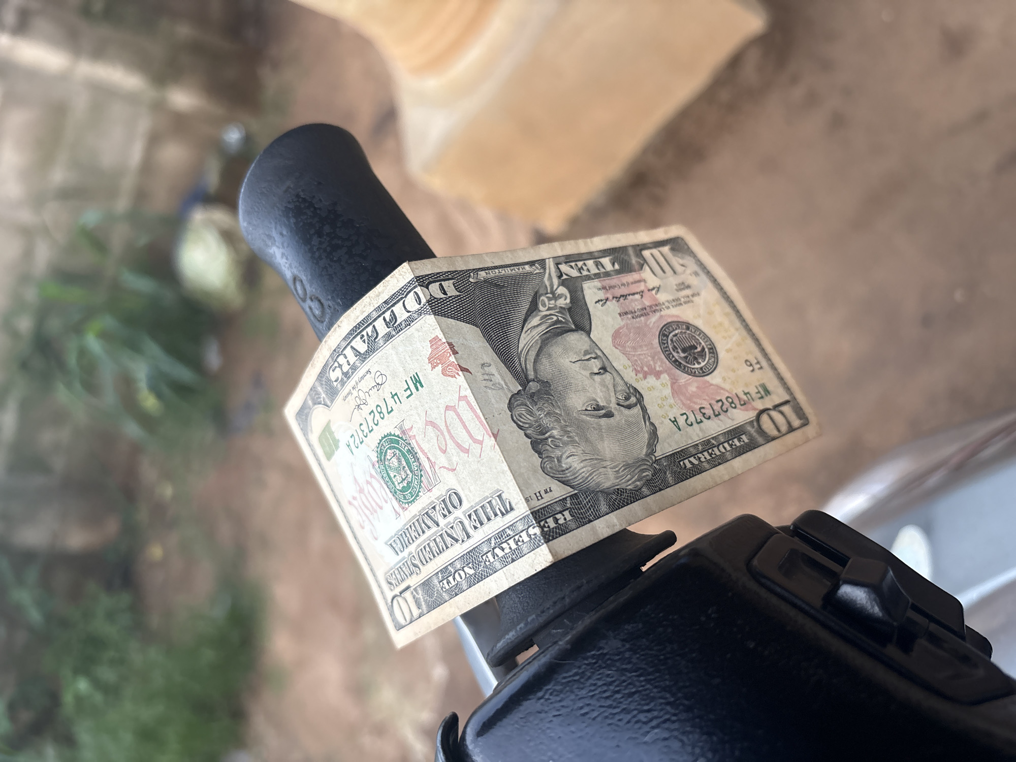 A close-up shot focuses on a partially obscured ten-dollar bill, appearing slightly crumpled and resting on top of what seems to be a black handlebar, possibly of a bicycle or motorcycle. The handlebar curves down out of the field of view. The background is blurred and indistinct, showing hints of greenery. The photograph is taken from a point-of-view above the handlebar, looking down at a slightly oblique angle. There is no information to suggest that this image was taken in Ikot Ekpene, Nigeria.