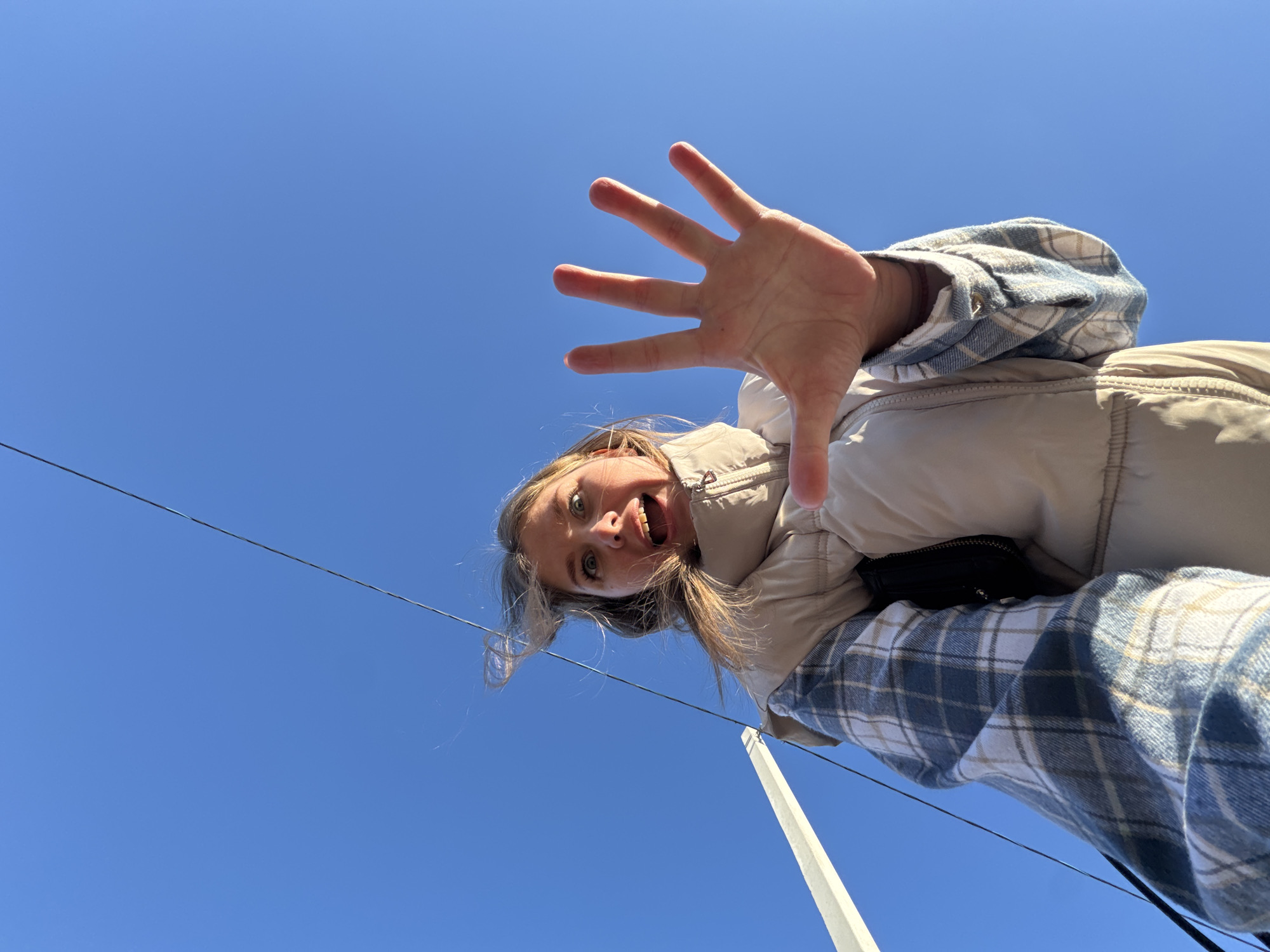 A low-angle, point-of-view shot depicts a woman outdoors against a clear blue sky. She has blonde hair, fair skin, and green eyes, and she is smiling with her mouth open. Her right hand is raised toward the camera, fingers splayed wide. She is wearing a light-colored padded jacket over a blue and white plaid shirt. A thin black wire stretches diagonally across the upper left of the frame, and a white pole rises vertically in the lower right. Based on the background, this location is not identifiable as the city of Горный, Russia, or any other specific location.