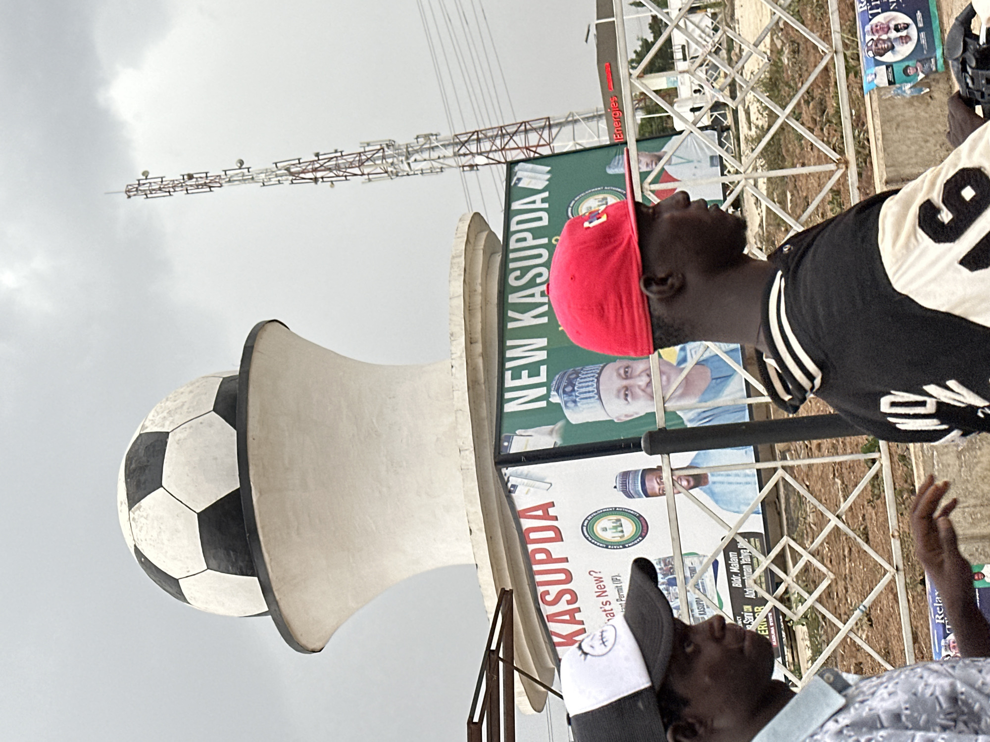 The image captures an outdoor scene, rotated 90 degrees clockwise, dominated by large promotional banners and structures under an overcast sky. In the foreground, a prominent white loudspeaker shaped like a megaphone, topped with a black and white soccer ball, is visible. Behind it, a man wearing a red cap and a black and white striped jersey (with a partial '9' on the back) looks intently upwards and to the right. Another man, wearing a white and grey cap, is visible in the lower left, also looking upwards.
Numerous large banners and billboards are erected on metal truss structures. These banners feature portraits of men, likely political figures or officials, in traditional attire. Visible text on these banners includes "KASUPDA," "NEW KASUPDA," and "What's New?," along with a partial phrase "ant Permit (IP)." One banner explicitly mentions "KADUNA STATE" and "Bldr. Malam Sani Abdurrahman Yahya Ph," suggesting the location is Kaduna State, Nigeria, and possibly related to a political campaign or public development initiative. Other fragments of text like "Energies," "Relax," "Ti," and "N" are also present.
In the background, a tall antenna or mast structure with multiple crossbeams extends into the sky, along with various wires. The overall impression is of a public event or gathering, possibly a political rally or community announcement, given the large loudspeaker, banners, and the attentive gaze of the visible individuals. The time of day appears to be daytime, under cloudy conditions.