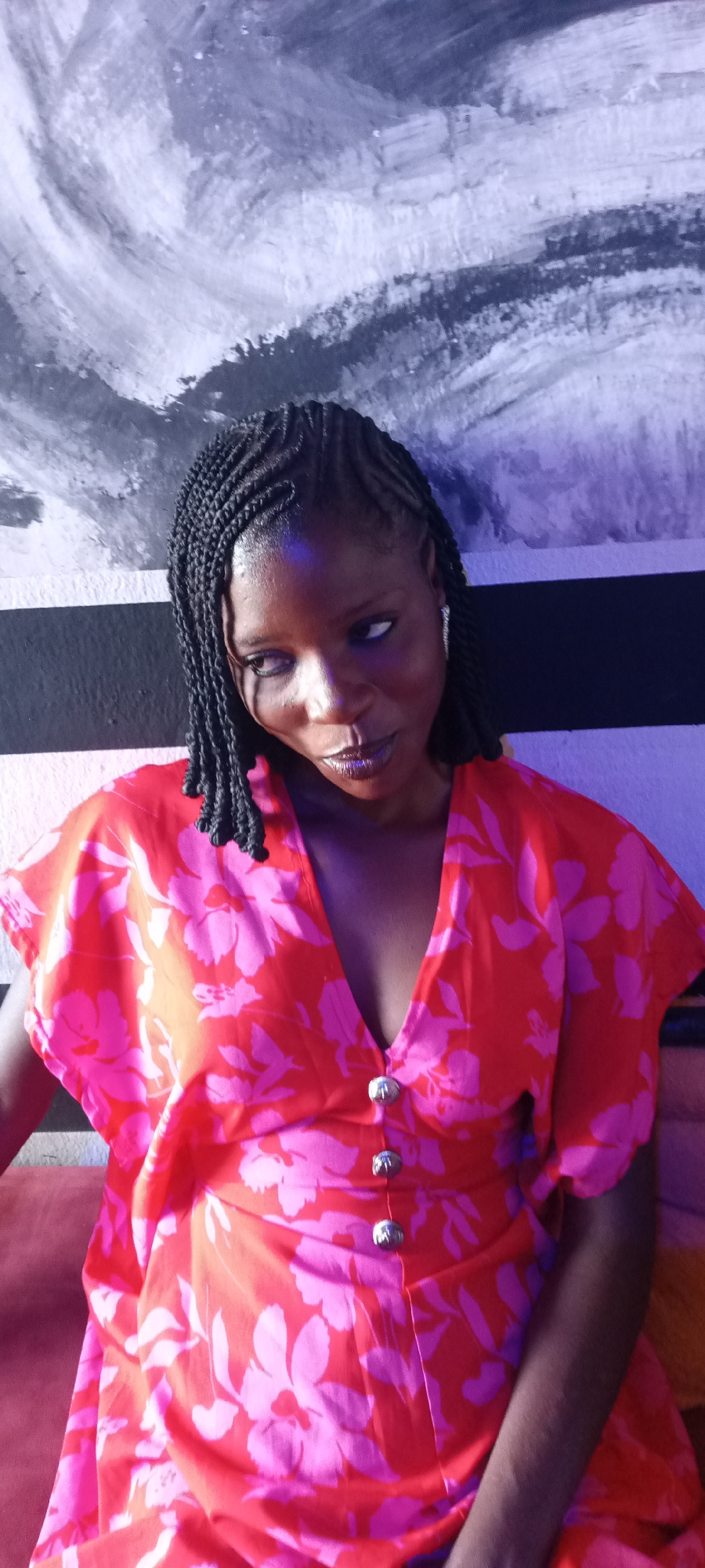 A woman is photographed indoors, likely in Jos, Nigeria, evidenced by her style and features, with a distinctive backdrop featuring horizontal stripes of black and white, overlain with an abstract design in shades of grey and white. The woman has dark skin, braided hair, and is wearing a red dress patterned with pink floral designs, fastened by three silver buttons at the chest. Her gaze is directed towards her left, and she appears to be sitting or reclining on a soft, reddish surface. The scene is illuminated by a light source that casts a purplish hue onto the backdrop and parts of the woman's face.