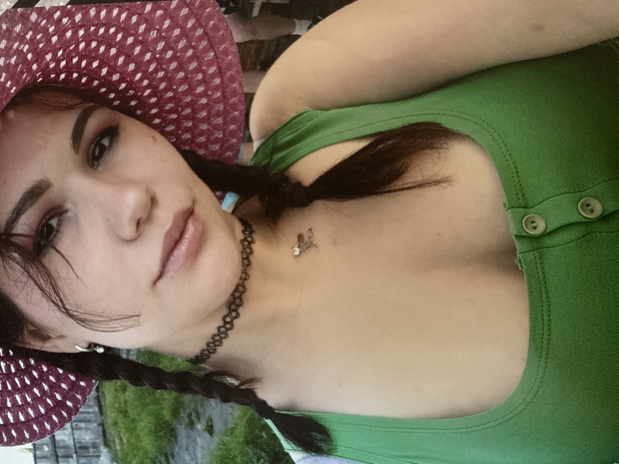 A young woman wearing a red and white polka dot hat, a black choker necklace, and a green tank top is shown in a close-up selfie. She has dark brown hair styled in two braids, and she is wearing subtle eye makeup. A small pendant necklace hangs around her neck. The background appears to be an outdoor setting with greenery and a wooden fence. There is no clear indication of the city of Cheboksary, Russia, or any specific location details beyond what is visible in the image.