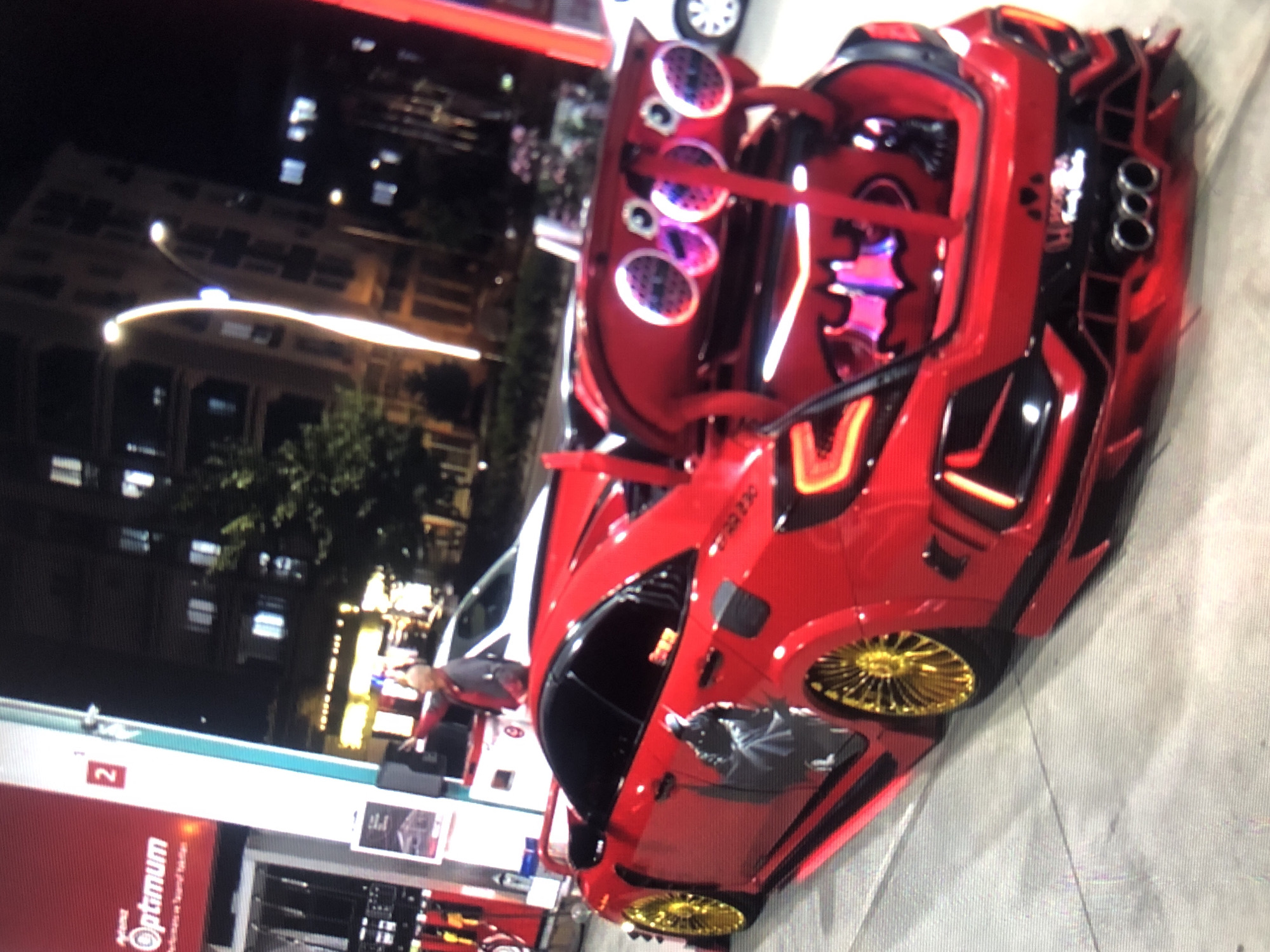 The image shows a heavily modified red car with its trunk open, revealing an elaborate sound system with multiple speakers illuminated by colorful lights. The car has custom bodywork and gold-colored wheels. The scene appears to be a gas station at night. An "Optimum" logo is visible on the gas pump. There is a building with illuminated windows in the background. The location is likely in Lhokseumawe, Indonesia. A person is standing near the car.
