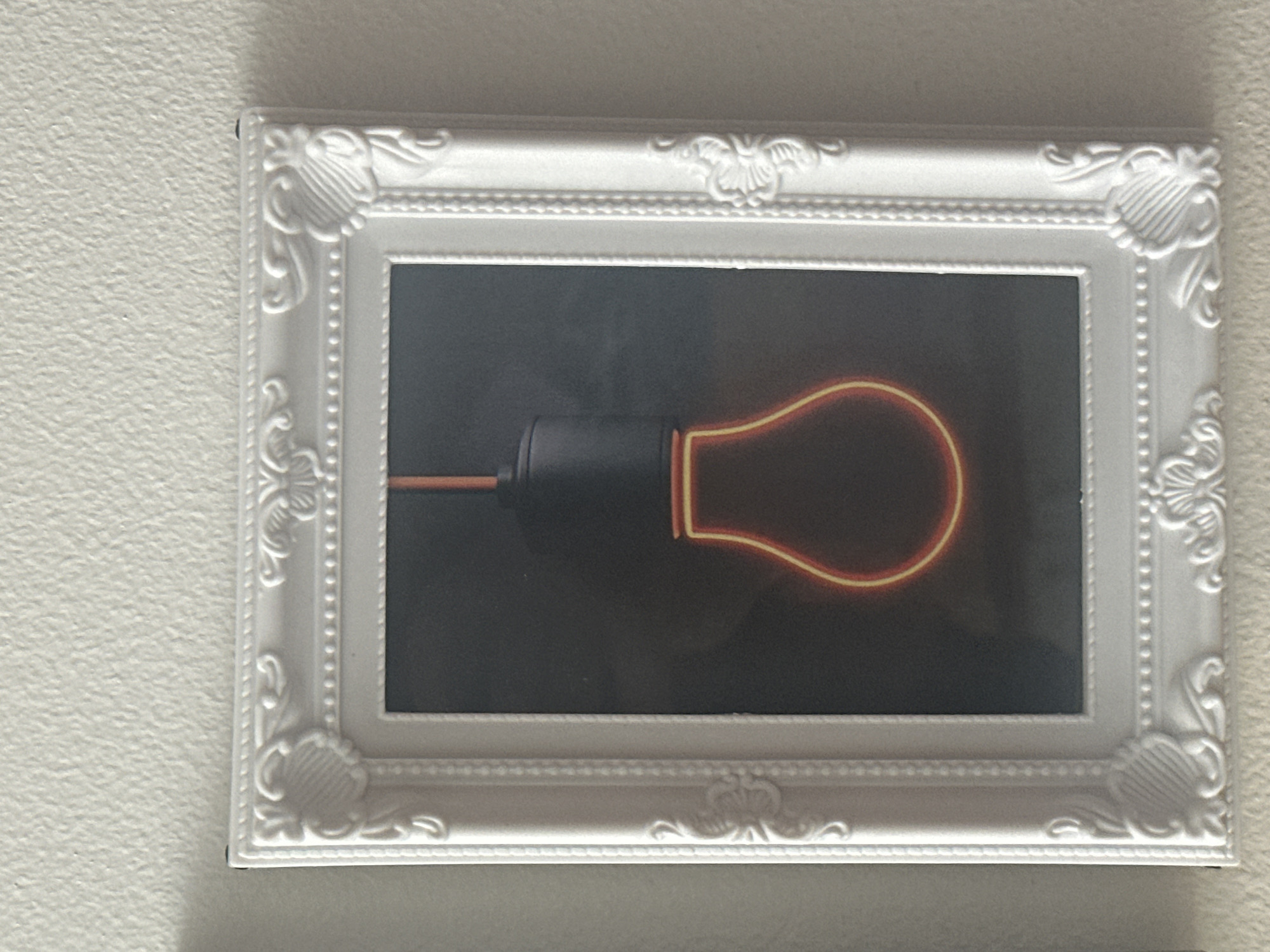 A framed picture of a neon light bulb hangs on a textured off-white wall. The frame is white with ornate carvings at each corner and along the edges. The picture inside the frame features a dark background with a stylized neon light bulb in orange and red. The bulb is suspended by a black cord. The scene is indoors, with the wall and picture being the sole focus. There are no people or other objects visible in the image.