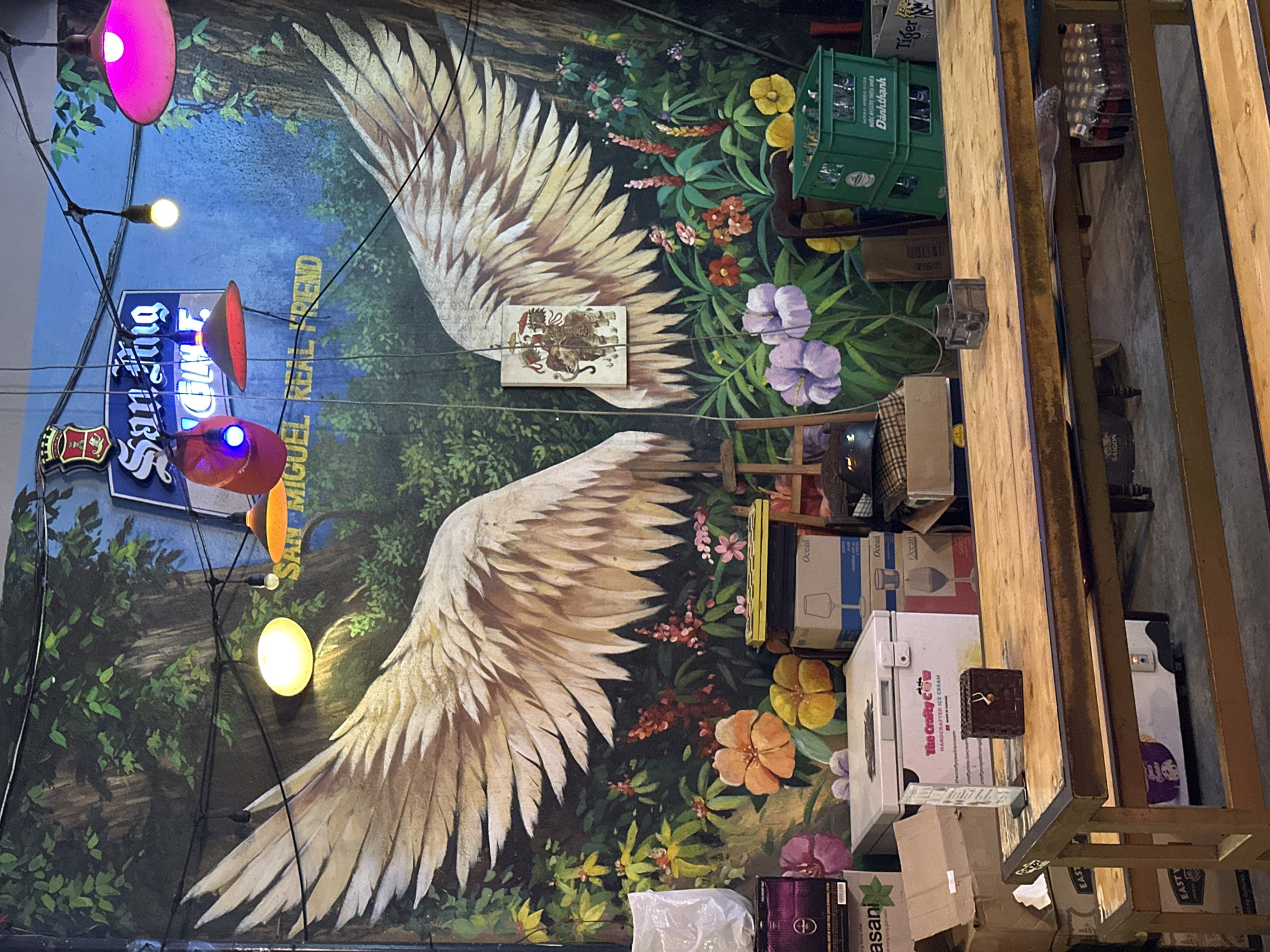 The image depicts a wall mural in what appears to be Đà Nẵng, Vietnam, given the presence of "Nước khoáng thiên nhiên Dành thành" (translated as Dành thành natural mineral water). The mural features large, stylized wings extending from a central image of the Mexican coat of arms, superimposed onto a background of dense foliage and vibrant flowers. "San Miguel Real Friend" is displayed in neon lettering. The area in front of the mural is cluttered with various objects, including boxes, crates of drinks ("Dành thành" and "Tiger Beer"), a mini-fridge branded "The Crafty Cow Handcrafted Ice Cream," and miscellaneous items like boxes labeled "Ocean" and "Easani." A wooden structure, seemingly a bar or shelf, is prominently featured, partially obscuring the mural. Overhead, lights with colored shades (pink, orange, and yellow) illuminate the scene. No people are visible in the frame.