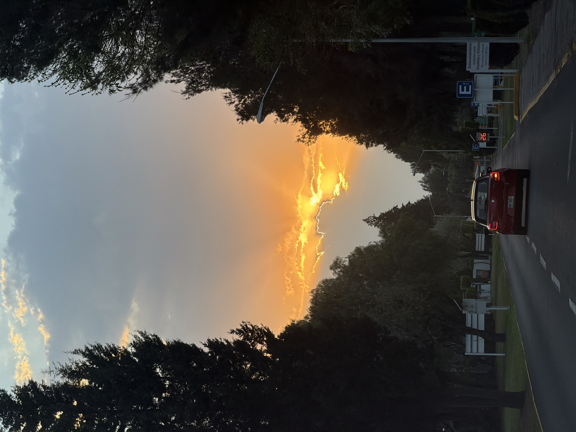 The image depicts a road in San Andrés Cholula, Mexico, framed by trees, leading toward a vibrant sunset. A red car, its license plate partially visible as 4SF-329, drives on the right side of the road. A sign indicates "Estacionamiento exclusivo para Estudiantes y Profesores de Lunes a Viernes 7:00 a 17:30 hrs," translating to "Parking exclusive for Students and Professors from Monday to Friday 7:00 to 17:30 hrs." The temperature display reads 26 degrees. A street lamp is visible on the left. The sunset casts an orange glow, partially obscured by trees. The sky also exhibits a gradient of grey and blue.