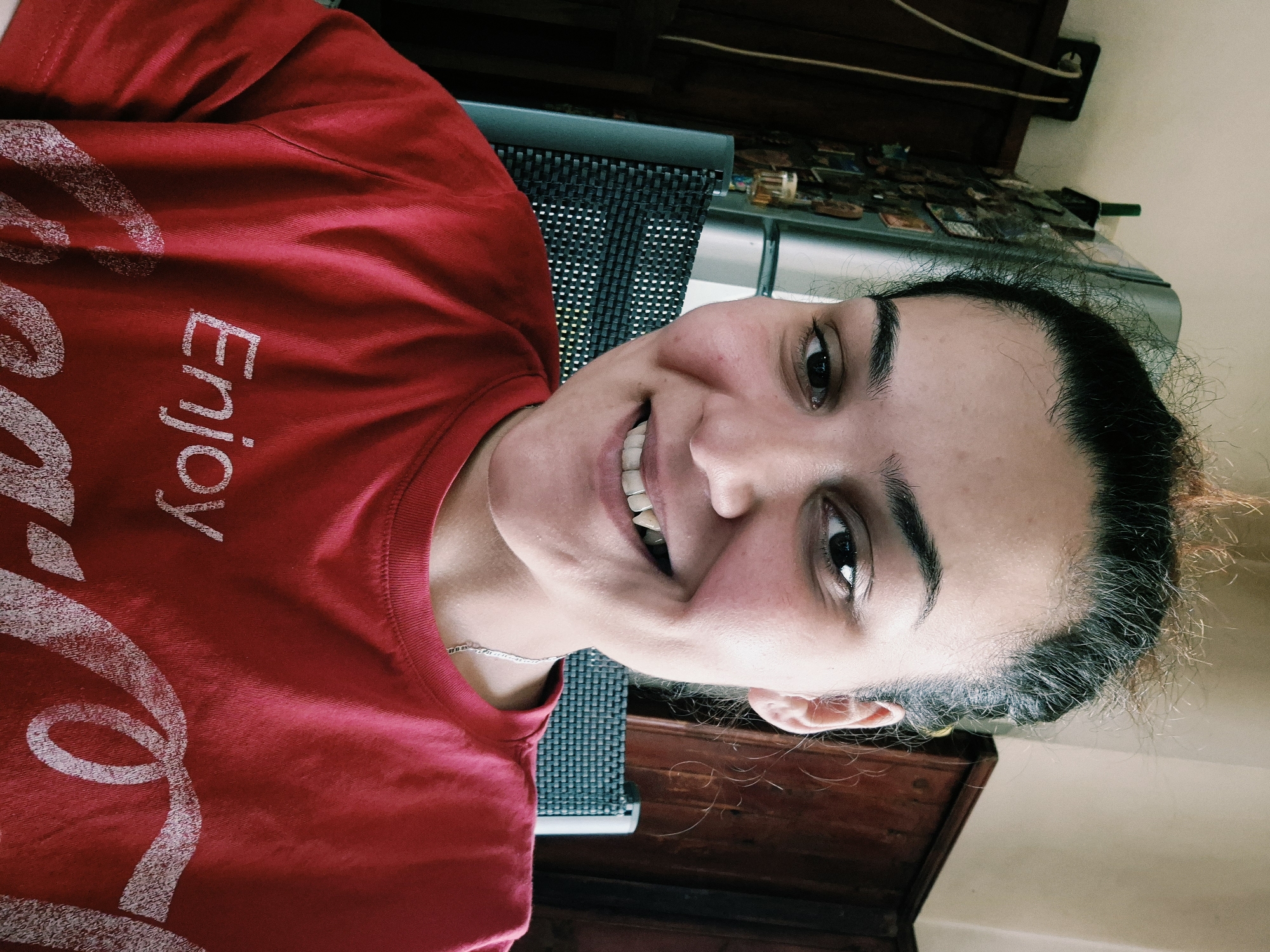 An adult female is depicted indoors in Iskrets, Bulgaria, in a medium shot, facing the camera directly. She is smiling, revealing her teeth, with a noticeable elongation of the upper right canine tooth. Her dark hair is pulled back, exhibiting numerous frizzy strands, some with lighter tips. Her eyes are dark, and both cheeks show areas of redness. She is wearing a red t-shirt with "Enjoy" and partially visible "Coca-Cola" branding printed in white text. A thin silver chain necklace is visible around her neck. The background includes a light-colored wall, dark wooden cabinetry or paneling, and the backrest of a dark grey woven chair positioned behind her left shoulder. To the left of the frame, a silver-colored refrigerator covered with various magnets is partially visible, with a white electrical outlet and two hanging cables mounted on the wall above it. A green object rests atop the refrigerator.