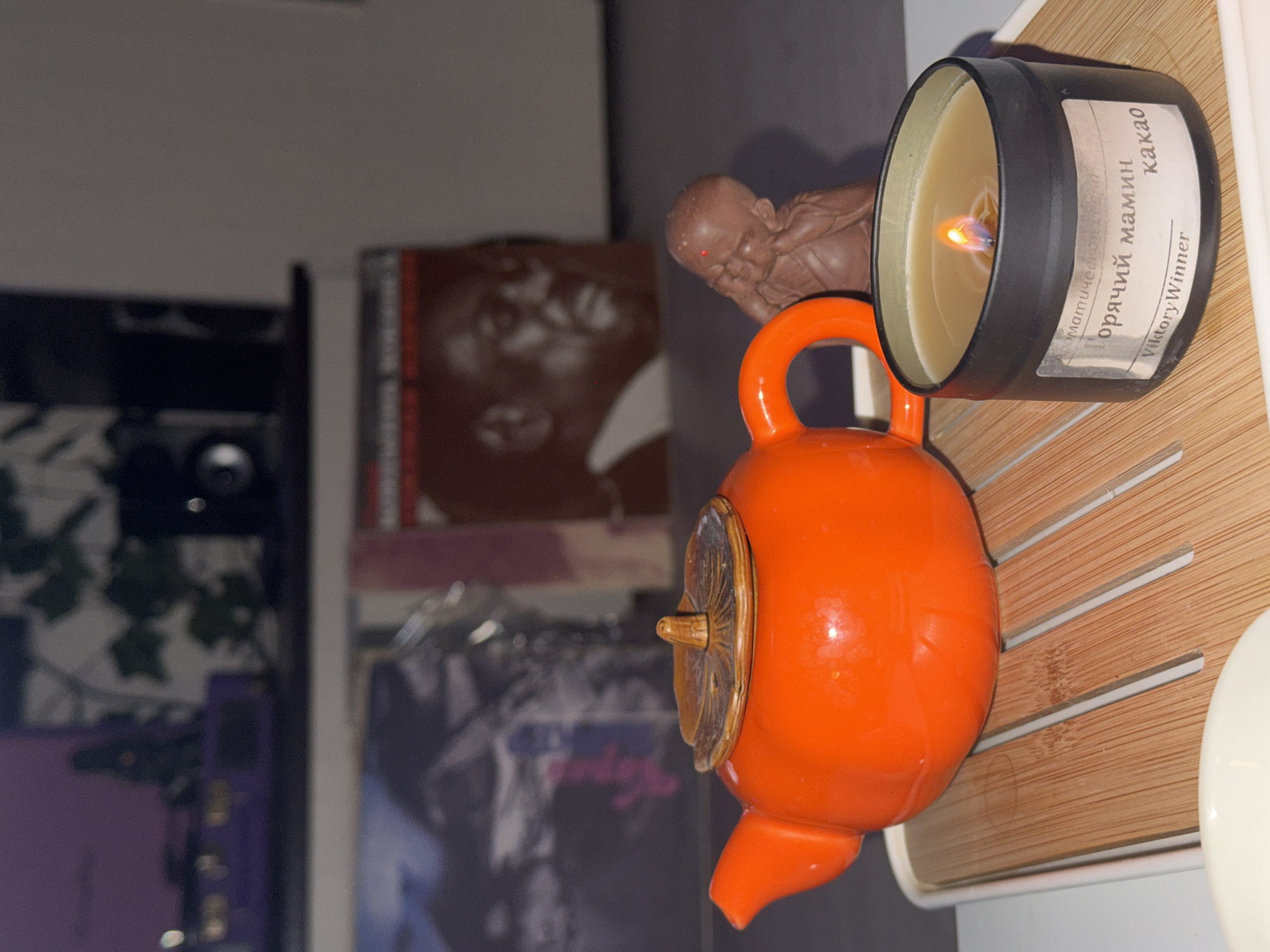 An orange teapot is placed next to a lit black candle with Cyrillic text on its label, and a small figurine. The objects rest on a slatted wooden surface. In the background, partially obscured by the teapot, are a record album, and other items, suggesting an interior setting. The city name Ново-Никольское, Russia is irrelevant since this is a photograph of objects and their surroundings.