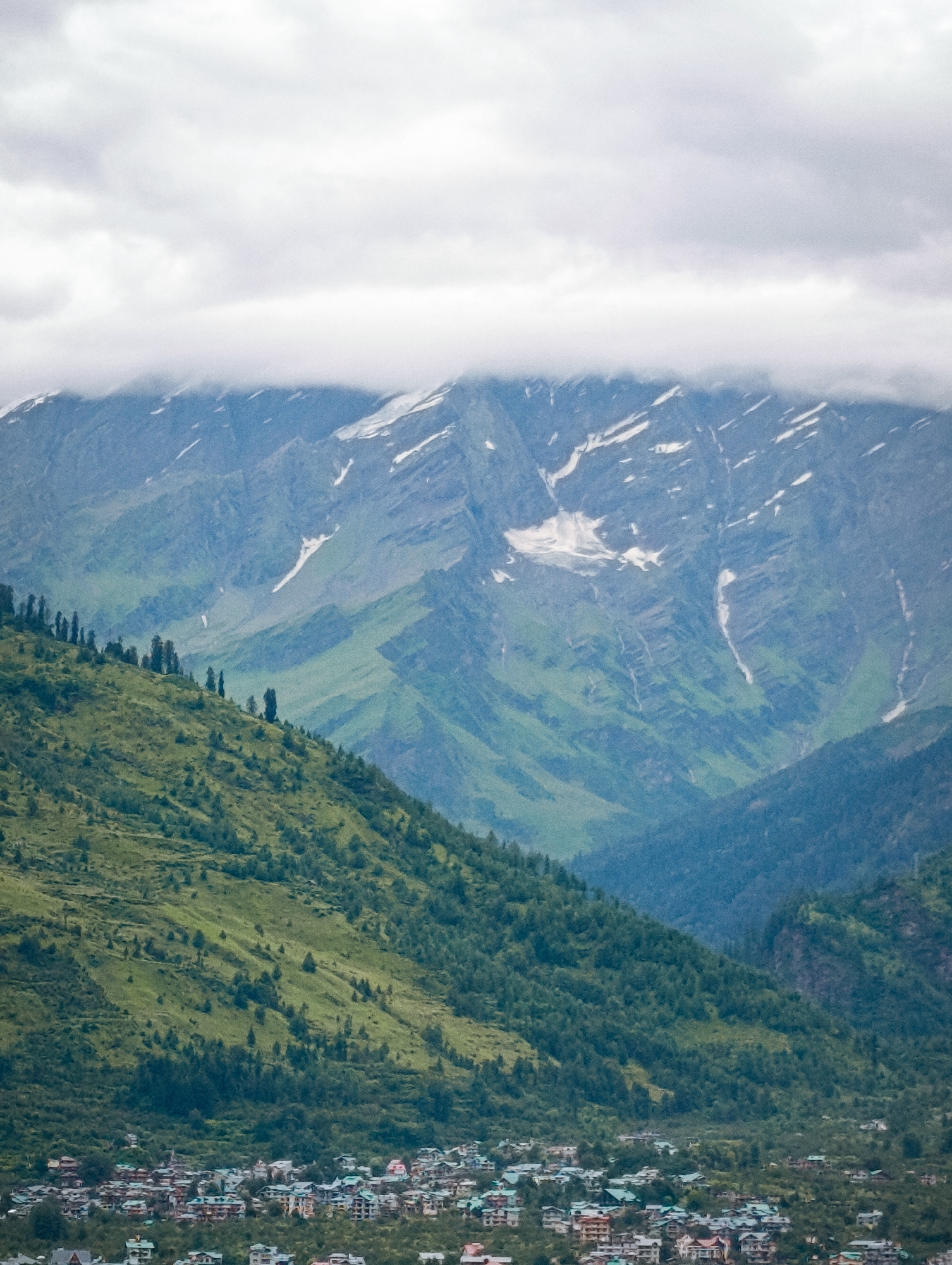 The image captures a panoramic view of a mountainous landscape, likely taken from a vantage point above the town of Vashisht, India. The foreground is dominated by a steep, verdant hillside dotted with scattered trees. Below this hillside, nestled in the valley, lies the town of Vashisht, characterized by a dense cluster of buildings with multicolored roofs. These buildings appear to be of varying sizes and architectural styles, indicating a built-up urban area. Beyond the town, the terrain rises sharply into rugged mountains, their peaks partially obscured by a thick layer of clouds. Patches of snow are visible on the higher slopes, suggesting a significant altitude. The overall atmosphere is one of natural grandeur, with the human settlement integrated into the dramatic mountain scenery.