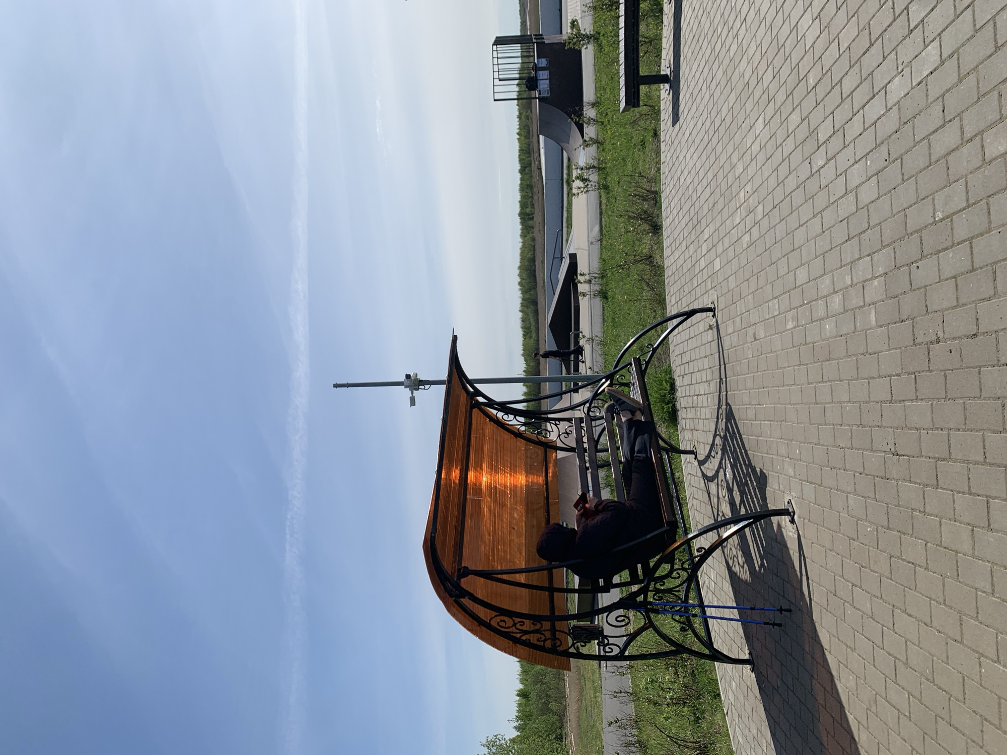 A photograph shows a person sitting in a metal outdoor bench on a paved walkway in Severodvinsk, Russia. The bench has a translucent brown roof, and is set in a grassy area near a body of water. The person appears to be looking at something in their hands, possibly a phone or book. Walking poles are visible near the bench. In the background, there is a small structure and more benches. The sky is blue with some wispy clouds. The paving stones are gray.