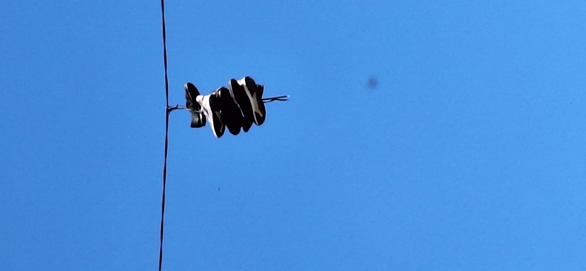 A pair of black and white athletic shoes hang from a black power line against a clear blue sky. A single power line runs vertically in the left of the frame with shoes looped and dangling. The location is most likely somewhere in a metropolitan area with power lines, but the scene lacks details that confirm the precise location in Blagoevgrad, Bulgaria.