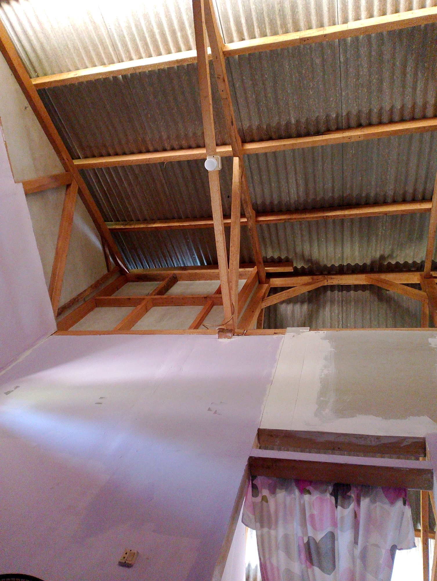 The image shows the interior of a partially constructed building in Desa Babakan, Indonesia. The ceiling is made of corrugated metal and supported by wooden beams. The walls are partially covered with a light-colored material, possibly drywall or plaster, with some areas unfinished. A white light fixture is visible on one of the wooden beams. In the foreground, there is a section of a wall painted a light purple. A light-colored electrical outlet is mounted on this wall. Below this wall, a curtain with a floral pattern is visible. The scene suggests ongoing construction or renovation work.