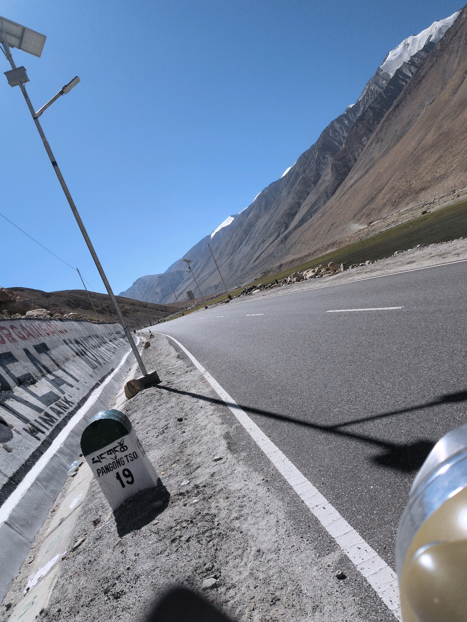 A perspective shot captures a mountainous landscape with a paved road winding through it under a clear blue sky. In the foreground, a roadside marker with Tibetan script and the text "PANGONG TSO 19" is visible, along with a solar-powered street light. The road is marked with white lines and casts a long shadow. The backdrop features towering mountains, some with snow-capped peaks, and a barren, rocky terrain. A concrete barrier with graffiti lines the left side of the road. The overall scene depicts a remote, high-altitude location.