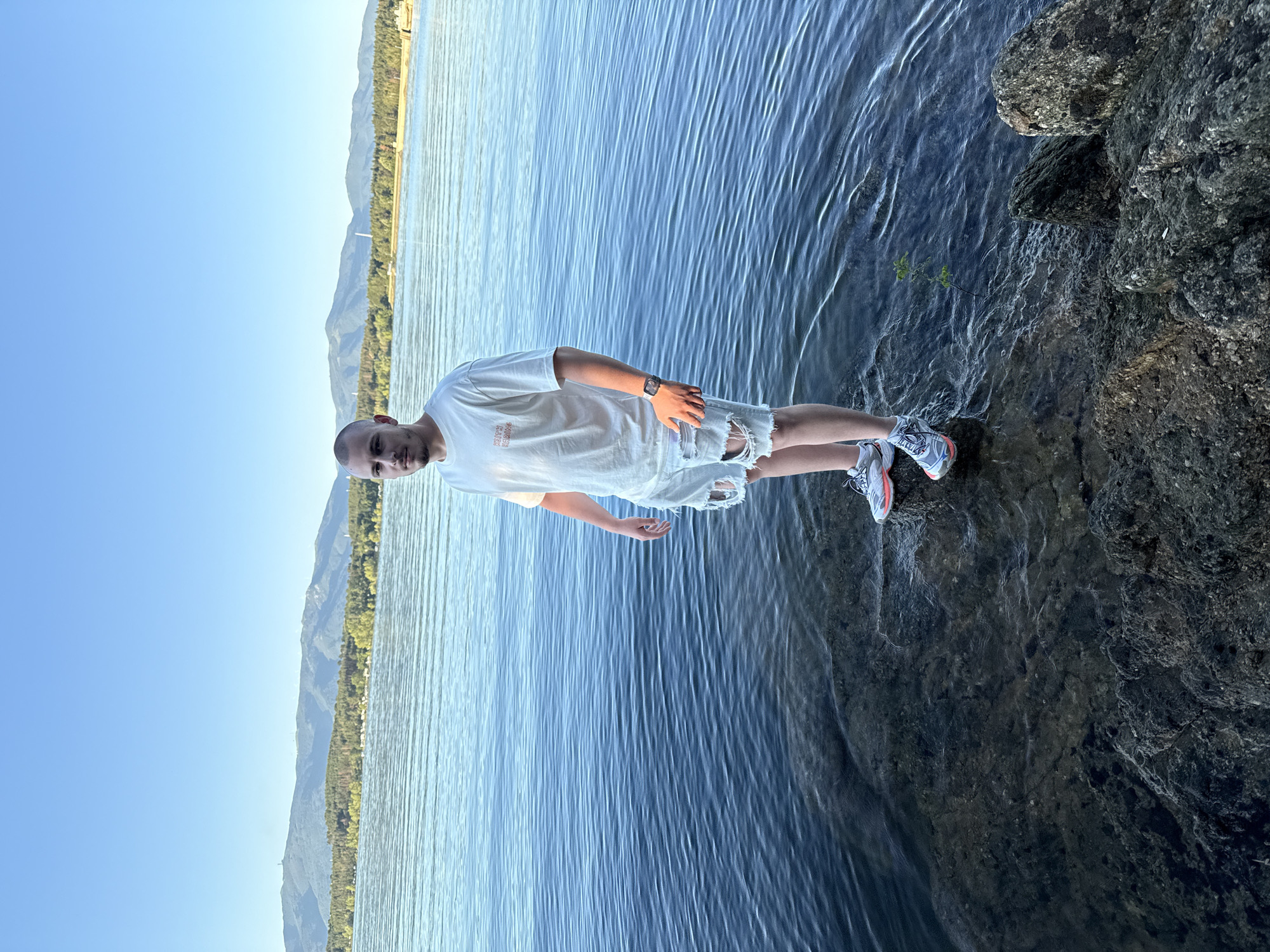 In Koprinka, Bulgaria, a man with a shaved head and a white t-shirt stands in the shallow water at the edge of the Koprinka Reservoir. He is wearing distressed denim shorts and athletic shoes. The water is calm, with small ripples reflecting the clear blue sky and distant mountains. The shoreline is rocky, with some larger boulders visible in the foreground. The scene is bathed in the warm light of what appears to be late afternoon or early evening.