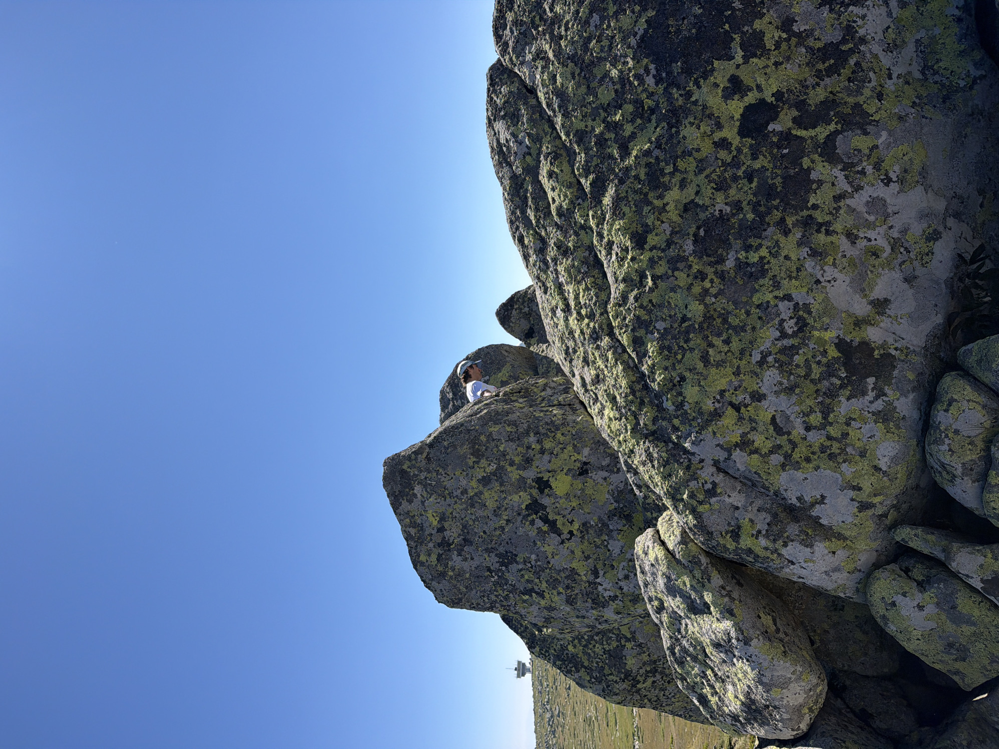 The image captures a natural, rocky landscape under a clear, bright blue sky. In the foreground, large boulders covered in patches of green and gray lichen dominate the scene. A person wearing a white shirt and a light-colored hat is partially visible, seemingly climbing or navigating among the rocks. In the background, a man-made structure with a tower and a dish antenna stands on a hill in the distance, suggesting a remote or elevated location. The ground around the boulders appears to be covered in sparse, dry vegetation. The overall impression is of an outdoor adventure or exploration in a mountainous or high-altitude environment. The mention of Bistritsa, Bulgaria, in the prompt suggests this location might be in or near that region, though the image itself does not contain any explicit urban elements or landmarks that definitively identify it as such.