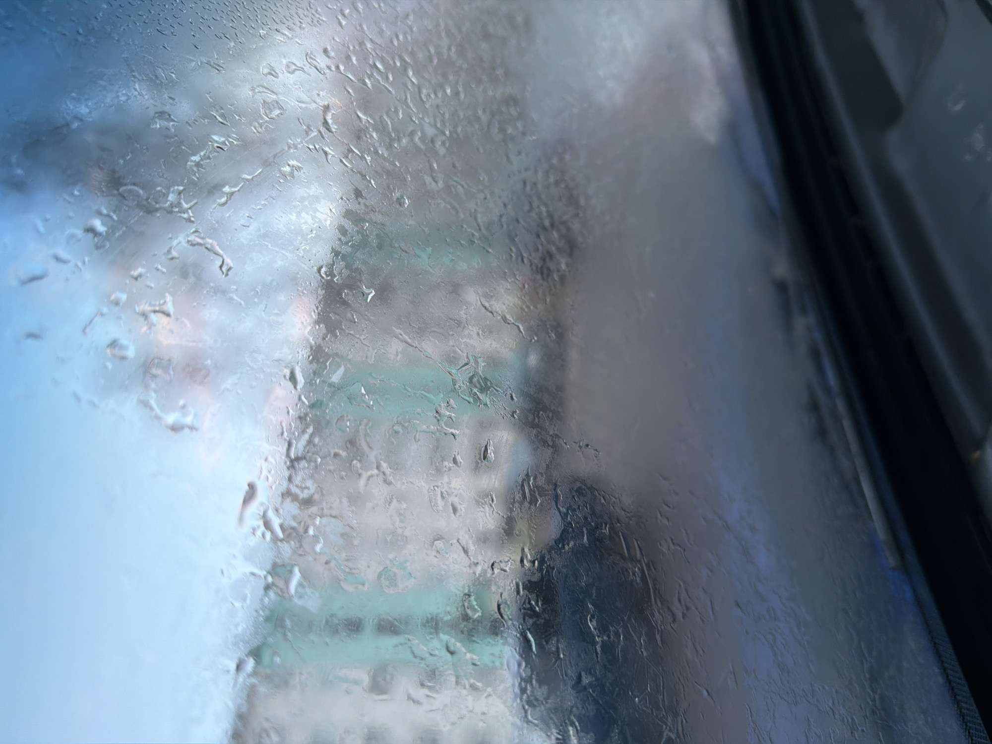 The image captures a view through a window heavily covered in water droplets and condensation, creating a blurred and abstract effect. The water distorts the external scene, which appears hazy and indistinct.
Through the watery surface, vague outlines and shapes can be perceived. There are suggestions of buildings or urban structures in the background, rendered in soft grays and muted teal tones. A dark, ambiguous silhouette, possibly a person or a large object, is faintly visible behind the droplets in the central-right portion of the image. The left side of the frame is brighter, with lighter blue hues, potentially indicating a sky or an open area.
The right edge of the image shows a dark, solid structure, likely part of a window frame or the interior of a vehicle, suggesting the photograph was taken from inside. The numerous water droplets clearly indicate rain or very high humidity.
No specific activity or event is distinctly taking place due to the obstruction. The time of day appears to be daytime, but the light is diffused, hinting at an overcast sky or a period of low natural light. There are no identifiable landmarks or specific location cues to pinpoint Chaikovsky, Russia. No text is visible in the image. The overall impression is one of an atmospheric, obscured, and somewhat contemplative scene.