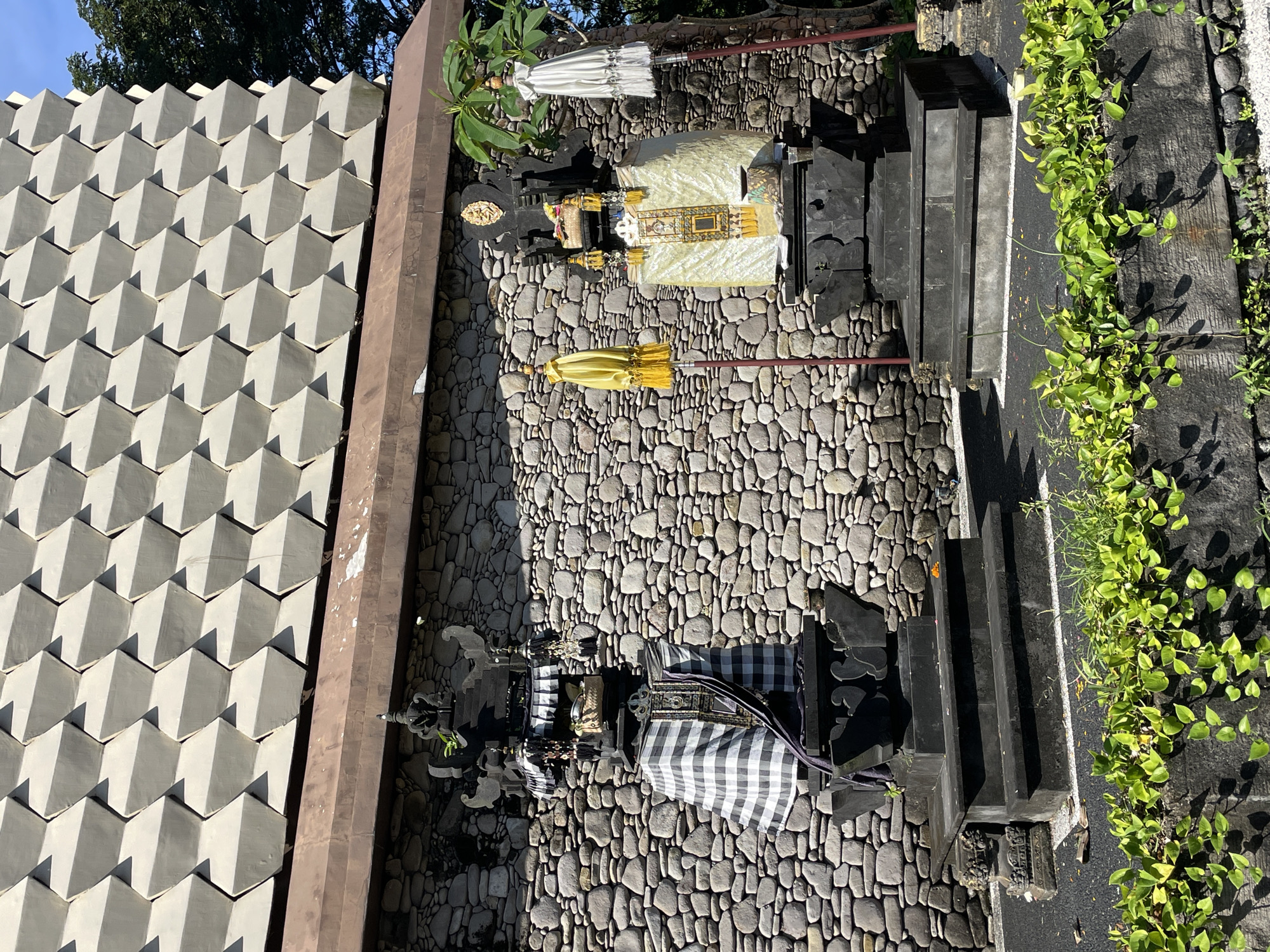 The image depicts a shrine in Kuta Selatan, Indonesia. The shrine is positioned on a ground paved with stones. It is ornamented with a black and white checkered cloth, as well as a yellow umbrella. Another similar structure, adorned with a lighter colored fabric and a white umbrella, is also visible. A building with a unique geometric, pyramid-like facade stands in the background. The building is grey and appears to be made of concrete. A brown post is present near the building. Vines and greenery are growing on the right side of the shrine. The sky is bright and clear, with visible tree branches at the top. The image is taken from an overhead angle.