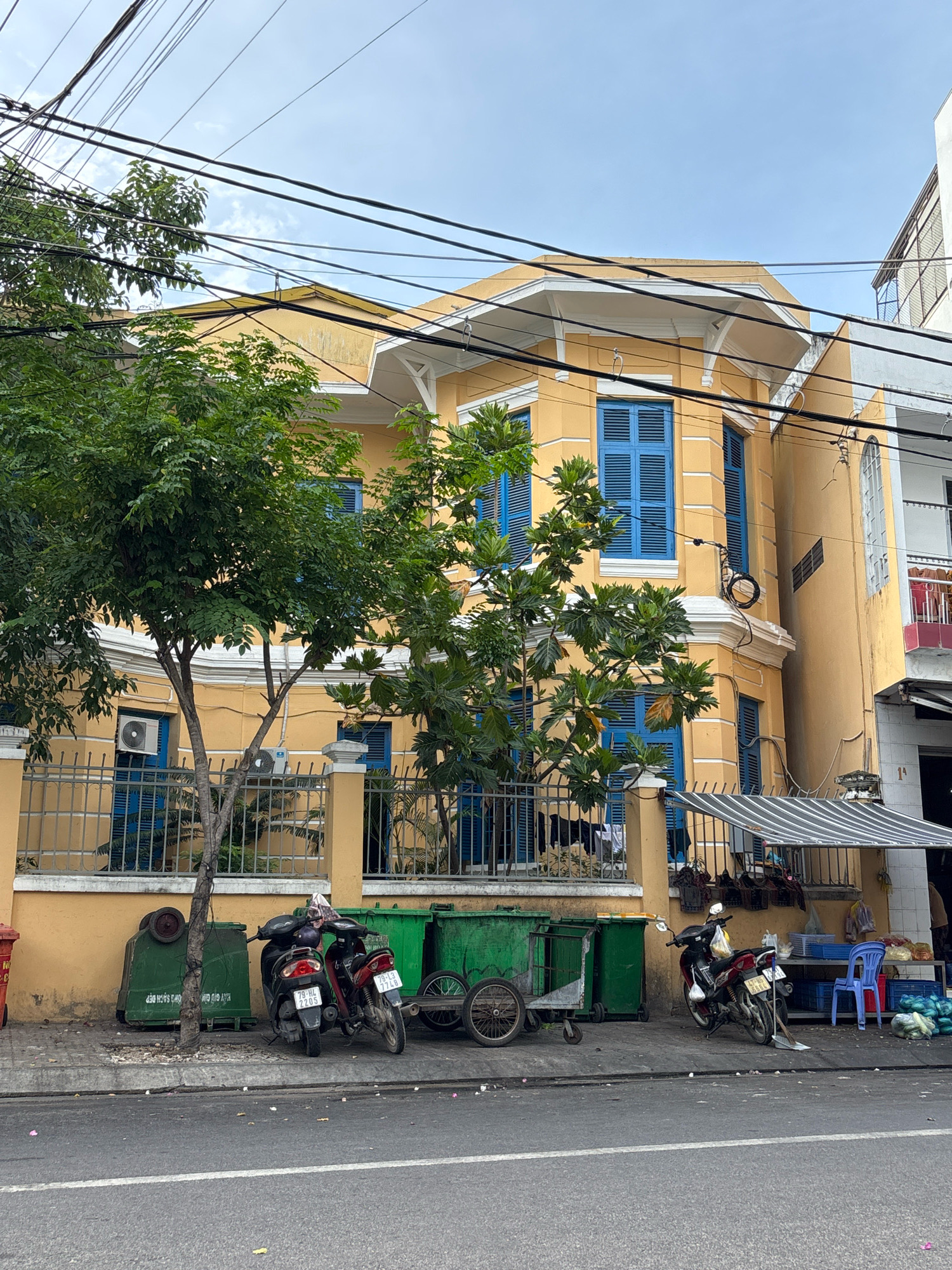 The image depicts a street scene in Nha Trang, Vietnam. The focal point is a two-story building painted in a vibrant yellow with blue shutters on the windows. The architecture suggests an older style, possibly colonial. In front of the building, a metal fence encloses a small yard with trees. Several green trash bins are lined up along the sidewalk, some appearing to be industrial-sized. Two motorcycles are parked on the sidewalk near the bins. The street in the foreground is paved and shows signs of wear, with debris scattered on the surface. Overhead, a complex network of electrical wires crisscrosses the sky. The sky is partially cloudy. To the right of the yellow building, another building with a white facade and a balcony is visible. An awning shades a small storefront area. Bags of produce, a blue plastic chair, and other items are visible near the storefront. The overall impression is a busy urban environment with everyday activities taking place on the street.
