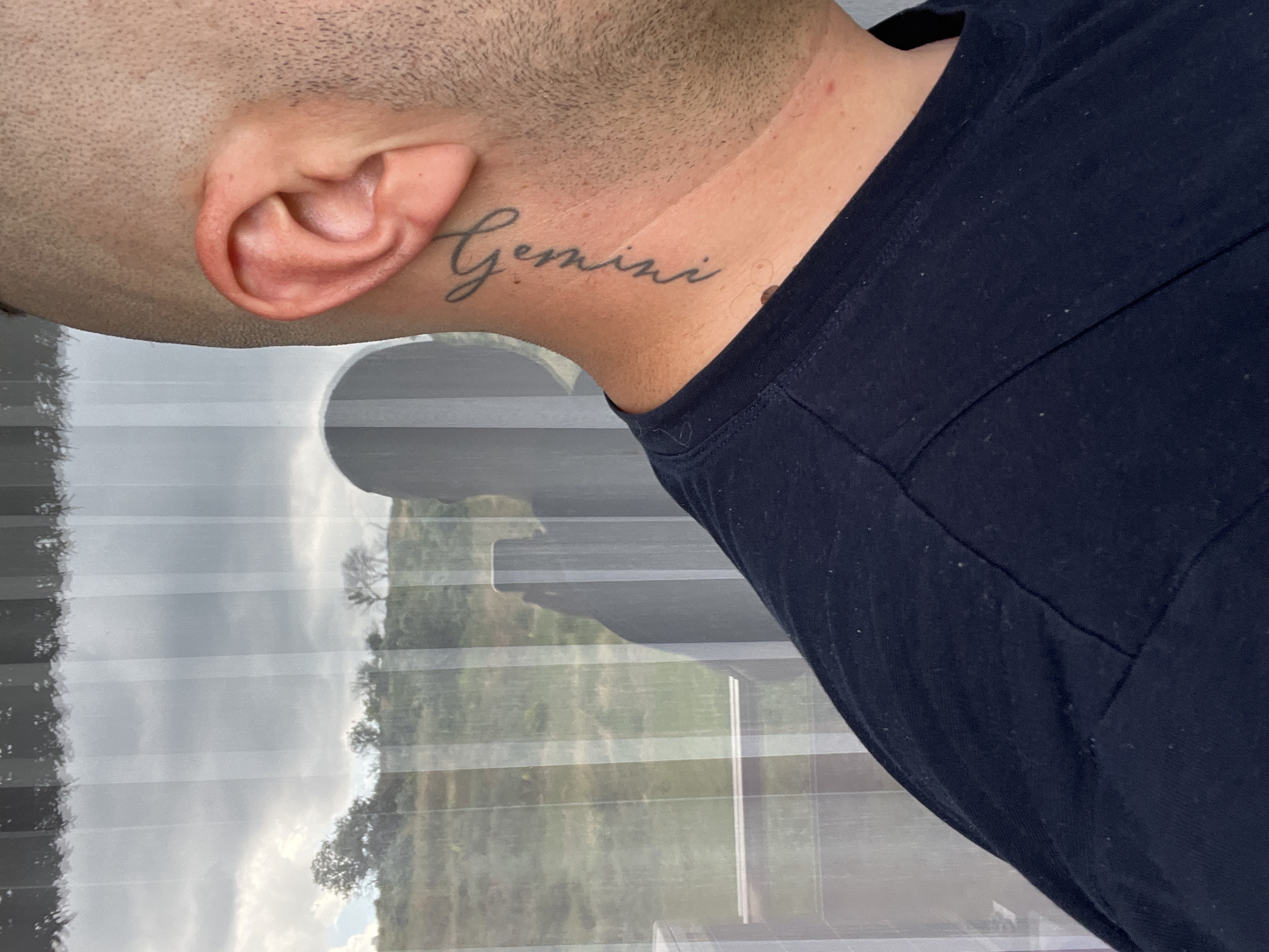 The image shows a close-up of a person's neck and ear, with a tattoo that reads "Gemini" on the neck. The person is wearing a dark blue t-shirt. In the background, a window shows a reflection of trees and the sky. There is no indication that this image is located in Varna, Bulgaria.