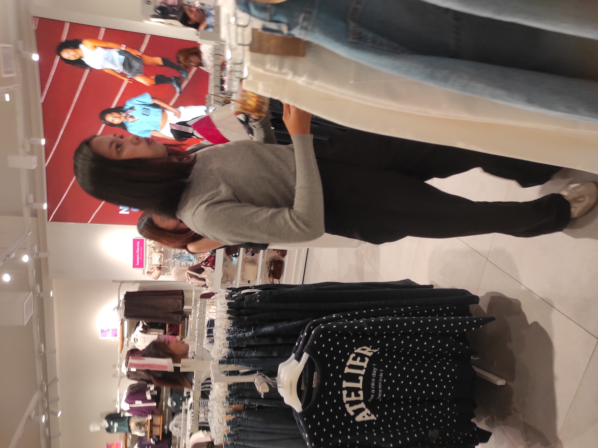 In a brightly lit clothing store, likely a fashion retailer, a young woman with dark hair and light skin, wearing a gray long-sleeved shirt and black pants, is the primary focus. She holds a clear plastic cup containing a beverage with ice and what appears to be liquid. She is standing in an aisle of clothing racks, examining items. In the background, a large red banner with images of three young women in athletic wear is displayed. Various other shoppers, some partially visible, browse merchandise in the background. Clothing items like jeans and sweaters are displayed on racks and shelves. The environment suggests a modern retail setting with white walls and ceiling lights. The floor is tiled.