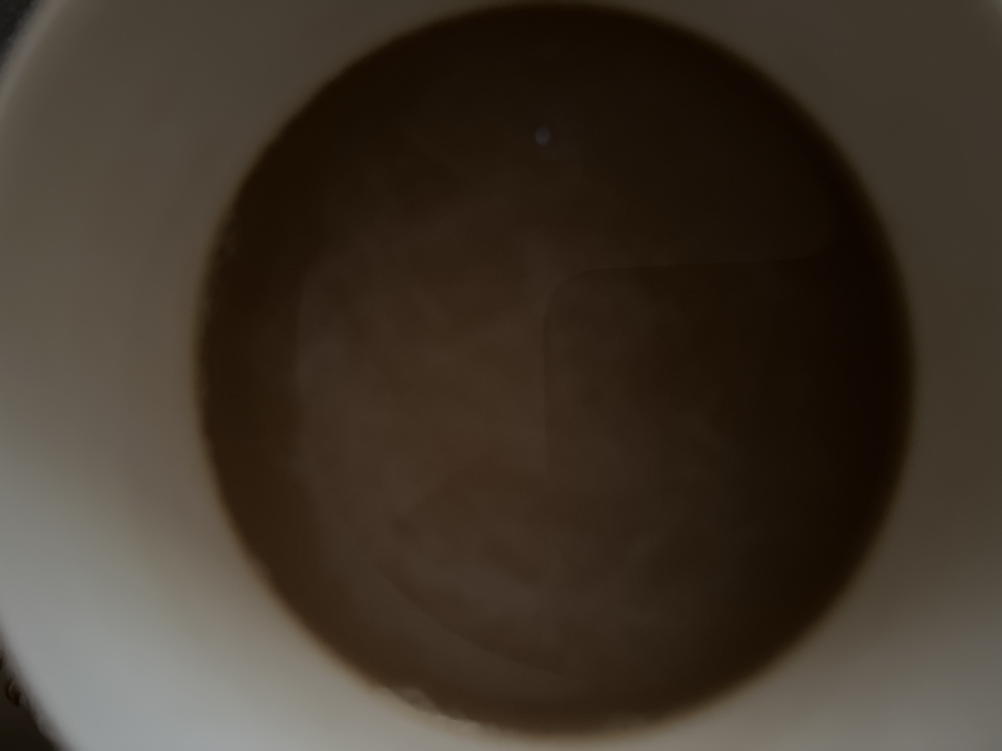 The image is a close-up, overhead view of a cup of coffee. The coffee is a medium brown color and appears to be steaming slightly. The rim of the white ceramic cup is visible around the edges of the coffee. There is a tiny, bright reflection visible in the coffee, possibly from a light source. The image is out of focus, giving it a soft, ethereal quality. There are no people or other objects visible in the frame. The scene depicts a simple, everyday moment.
It is not possible to determine the location in the city of Yerevan, Armenia, from this image.