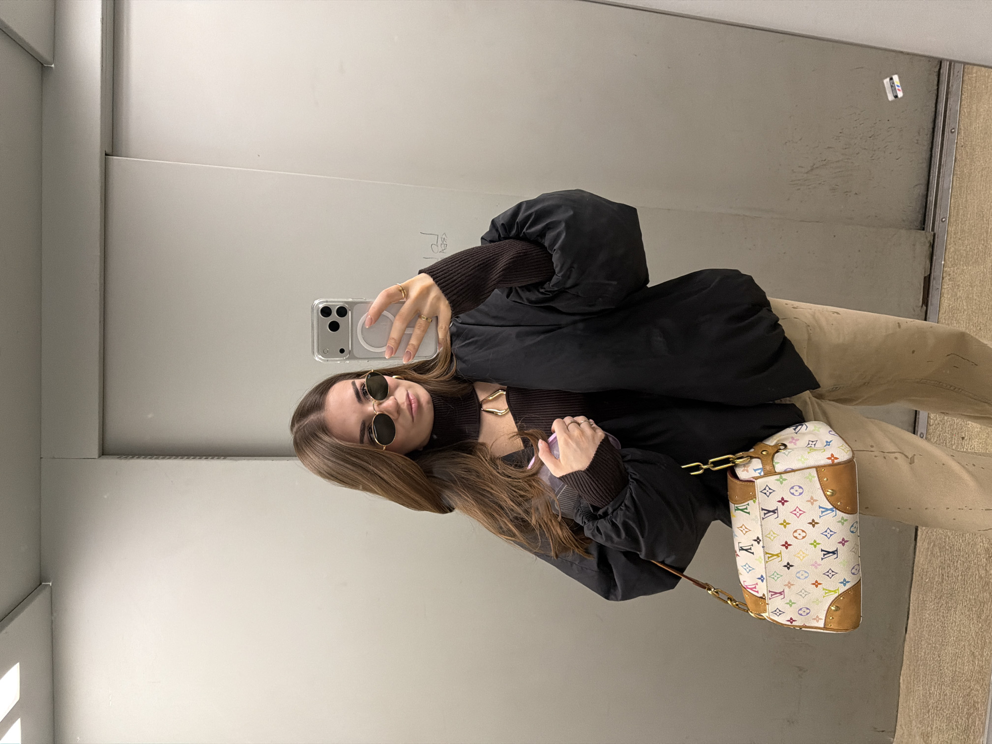 A woman is taking a selfie in what appears to be an enclosed space, possibly an elevator or a fitting room. She is holding her phone in her left hand, and it is in a clear case. The woman is wearing a brown turtleneck sweater under a black puffer jacket. She has on light brown pants. She has round sunglasses on and is wearing rings on her left hand. Hanging on her right side is a white Louis Vuitton trunk bag with colorful monogram detailing and gold hardware. The interior of the space is painted a neutral gray color. The location in Khimki, Russia is not supported by evidence in the image.